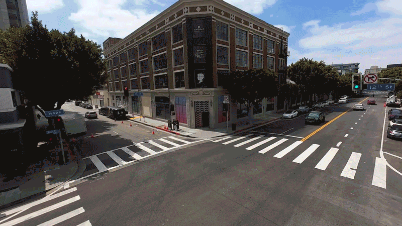 City intersection with crosswalks, traffic lights, and parked cars, in front of a multi-story brick building with storefronts.