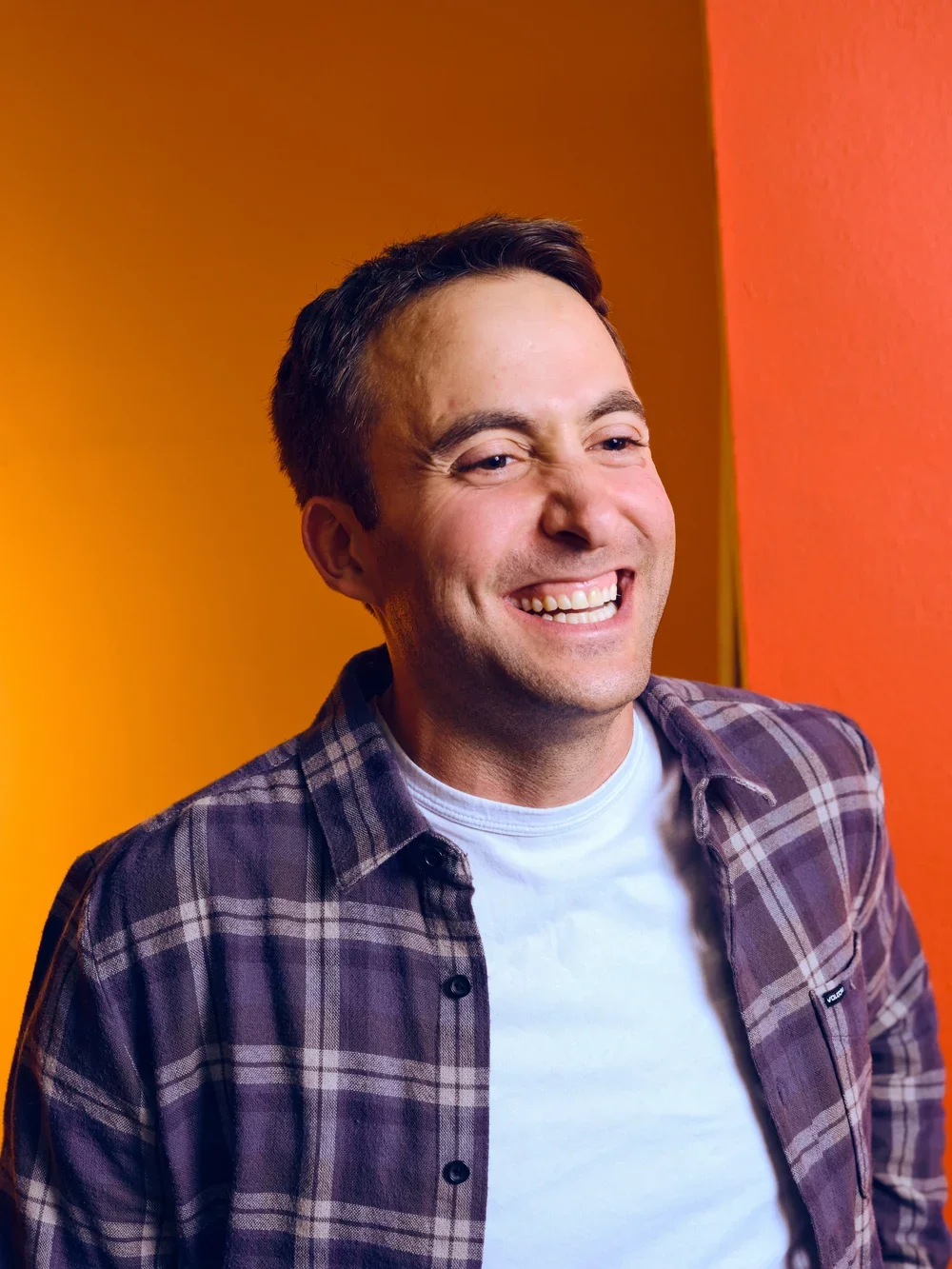 A man with slicked-back gray hair, wearing a blue short-sleeve sweater and beige pants, sitting on a cushioned bench against a plain light-colored wall with a shadow cast on it, looking to the side.