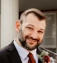 A man with a beard and short hair dressed in a suit, smiling outdoors near a building.