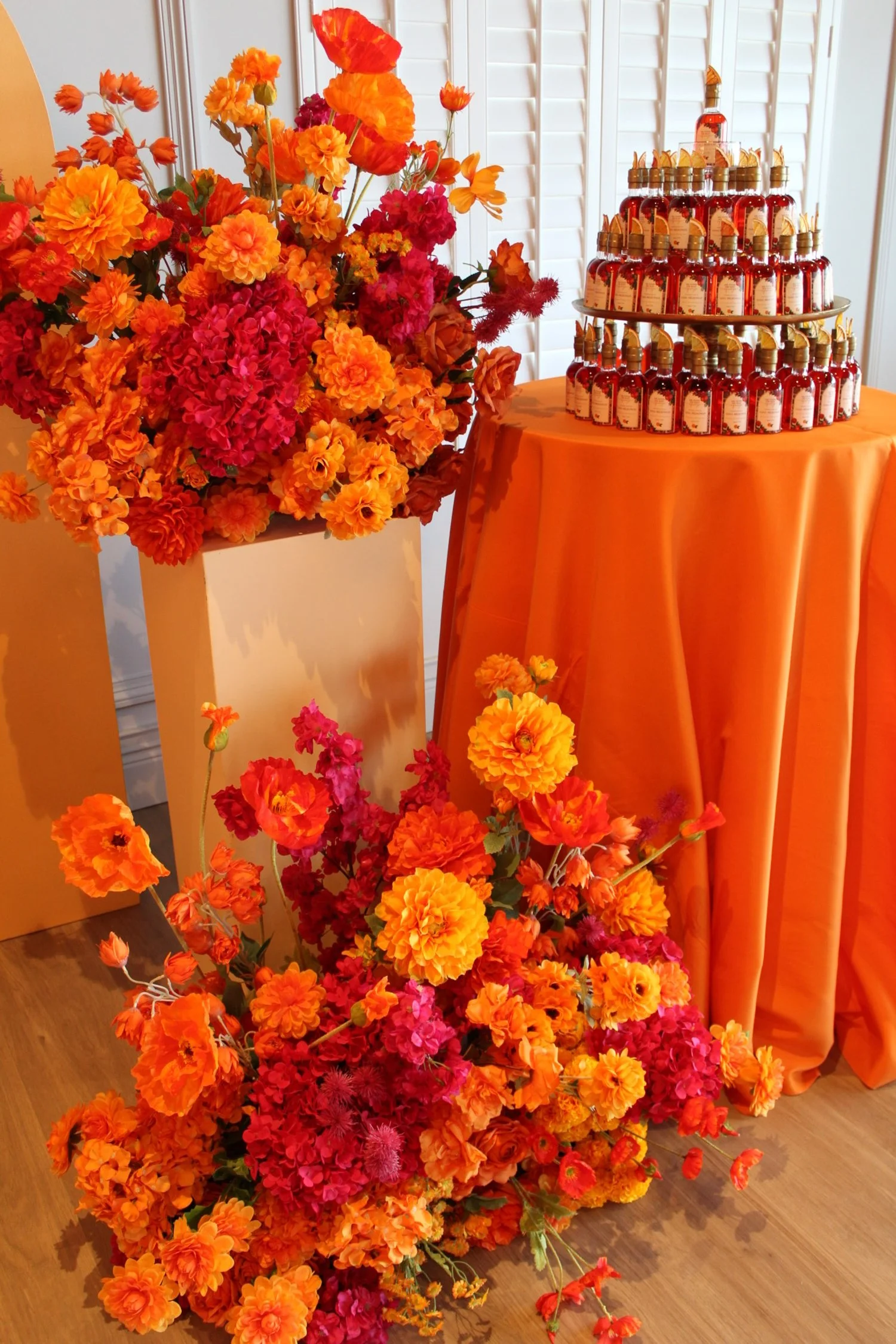 A vibrant floral display with orange, pink, and red flowers beside a table covered with an orange cloth. The table holds a tiered arrangement of small bottles with gold caps and labels. The backdrop features white plantation shutters.