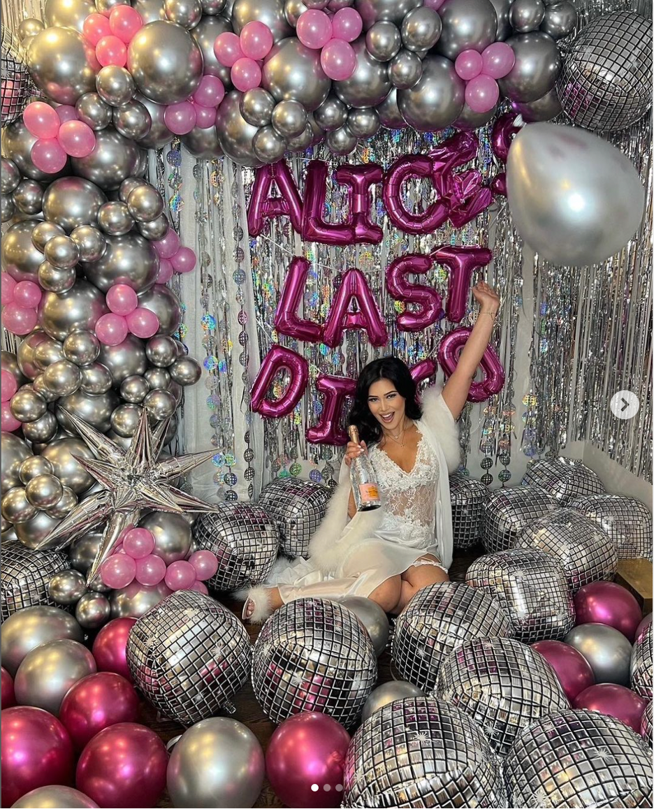 A woman celebrating her bachelorette party surrounded by pink and silver balloons, including metallic and disco ball styles, with pink foil balloons spelling out 'ALICE LAST DANCE'. She is sitting on the floor with a bottle of champagne, smiling and 