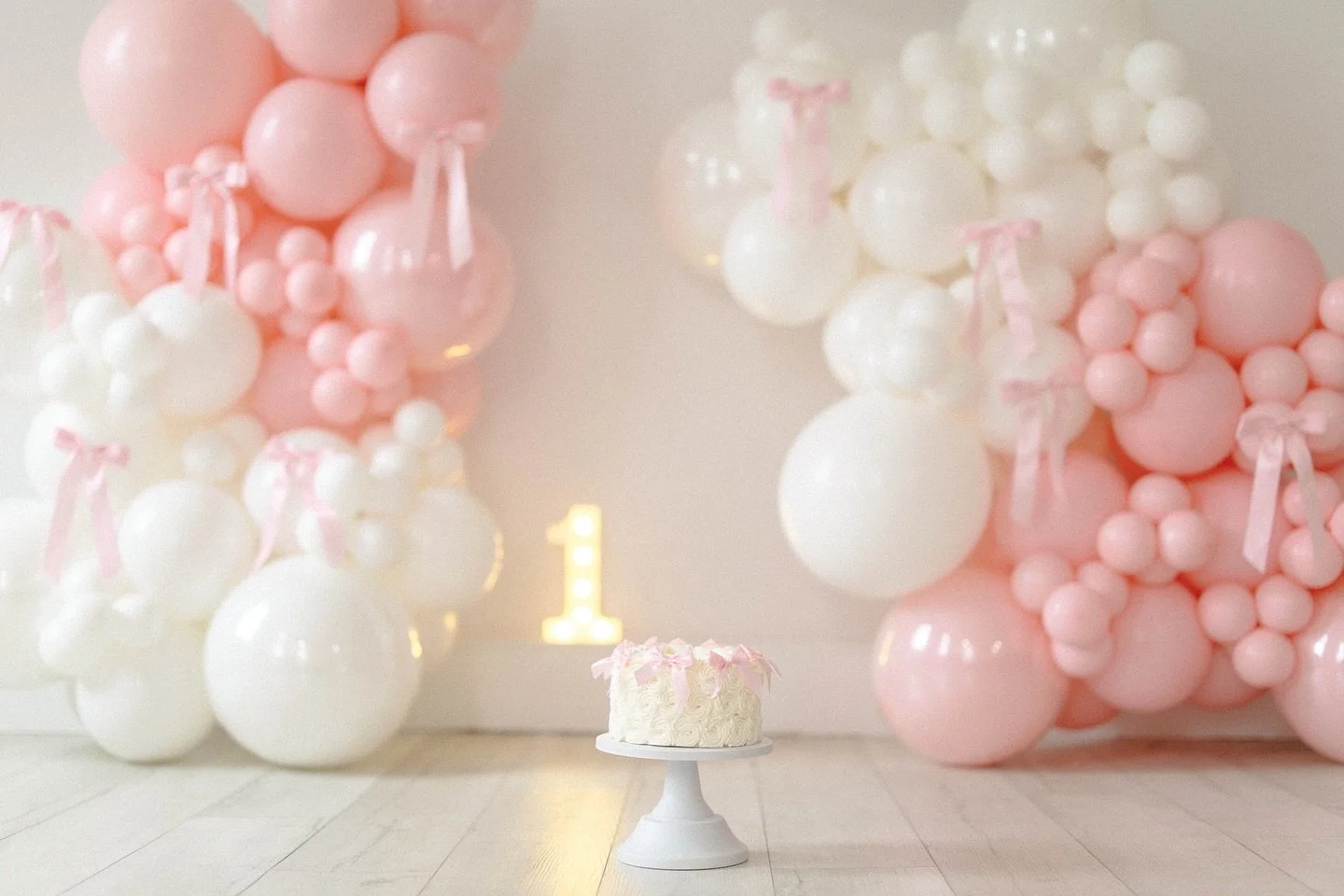 Celebration setup with white and pink balloon clusters and a cake on a white cake stand, with a lit number one sign in the background, indicating a first birthday celebration.