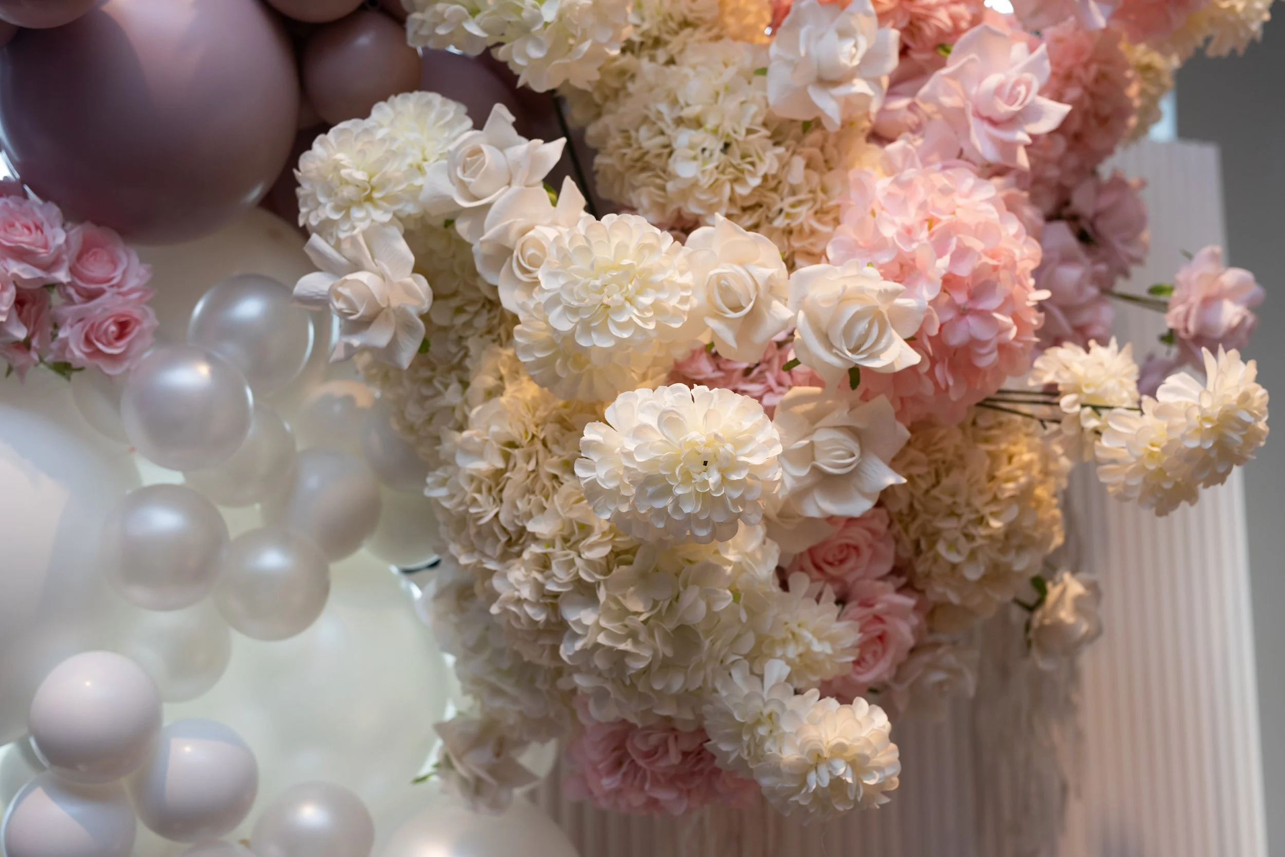 Arrangement of white, pink, and cream flowers with balloons in the background.