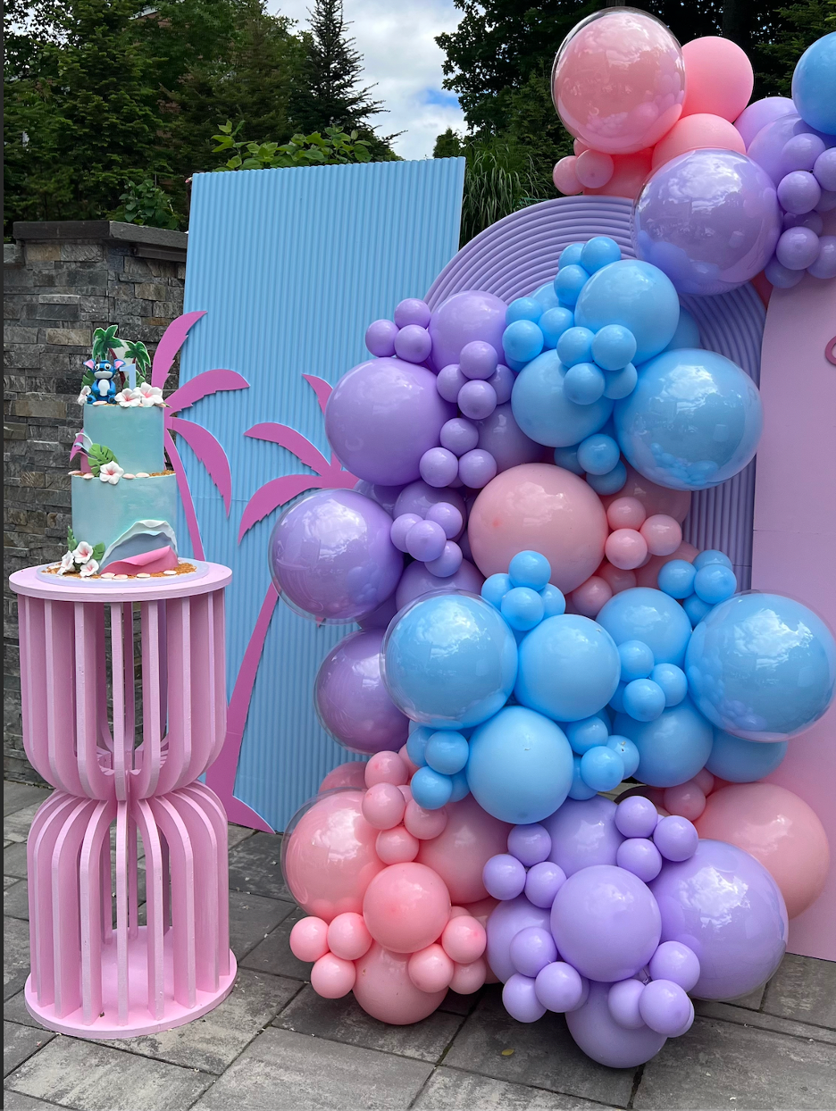 Decorative pink, purple, and blue balloons arranged with some large and small clusters, set against a blue and pink backdrop. A pink cylindrical stand holds a small cake decorated with tropical-themed elements like palm trees and a cartoon character,