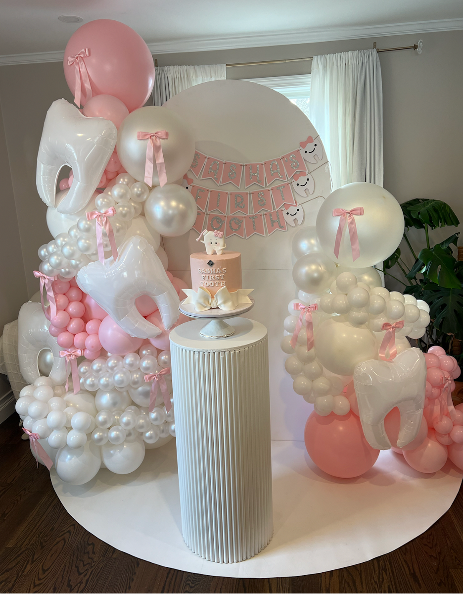 Decorative setup for a child's first tooth celebration with pink and white balloons, banners, and a small cake on a white pedestal.