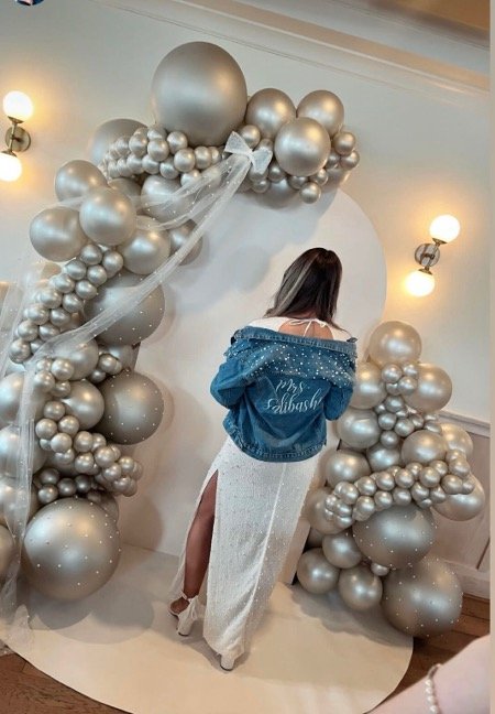 A woman stands with her back to the camera in front of a balloon arch made of silver balloons of various sizes, with a white sheer ribbon attached, in an indoor setting with warm lighting.