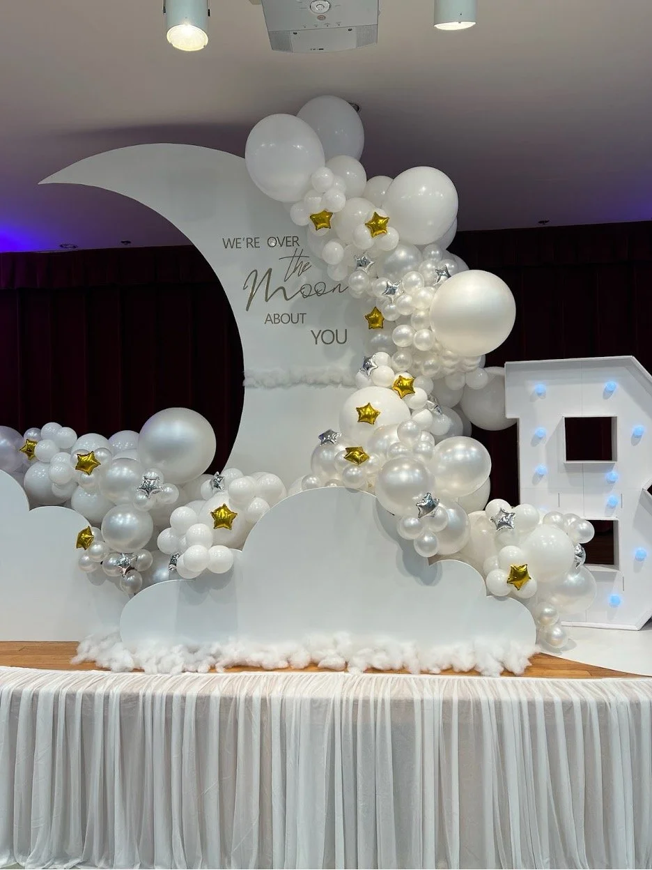 Decorative birthday or celebration setup with white clouds and balloons, a moon cutout with the text 'We're over the moon about you', and a large letter 'L' with blue lights.