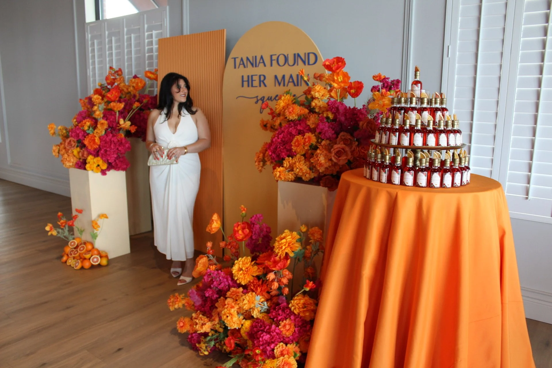 A woman in a white dress stands near a display of vibrant orange and pink flowers, with a sign that reads 'Tania found her man.' There is a table covered with an orange tablecloth holding numerous small bottles with dropper caps, arranged in a pyrami