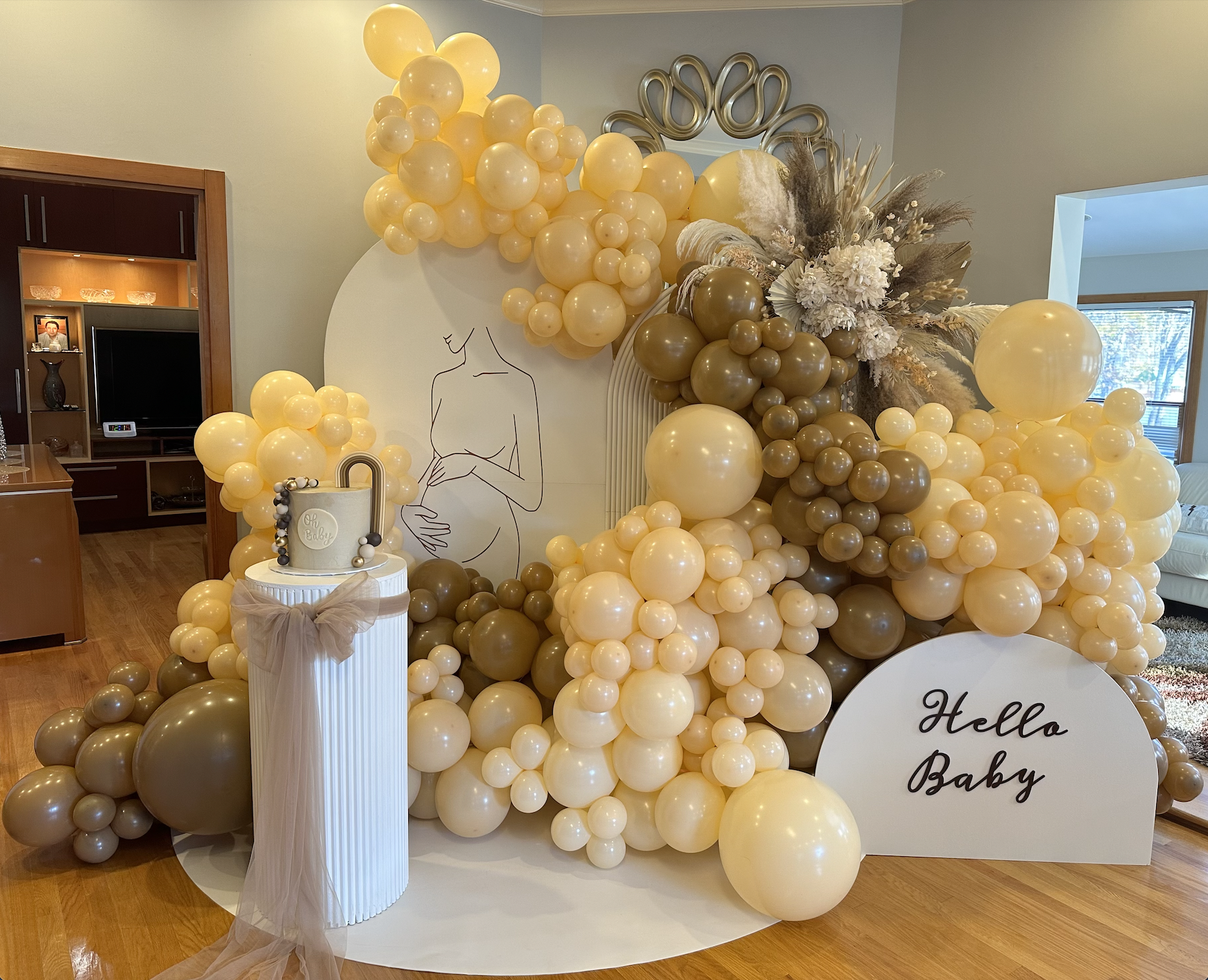 A decorated area with a cream and brown balloon arrangement, a white sign reading 'Hello Baby', a cake with a lock design on top, a minimalist line drawing of a pregnant woman on a white backdrop, and floral accents, likely for a baby shower celebrat