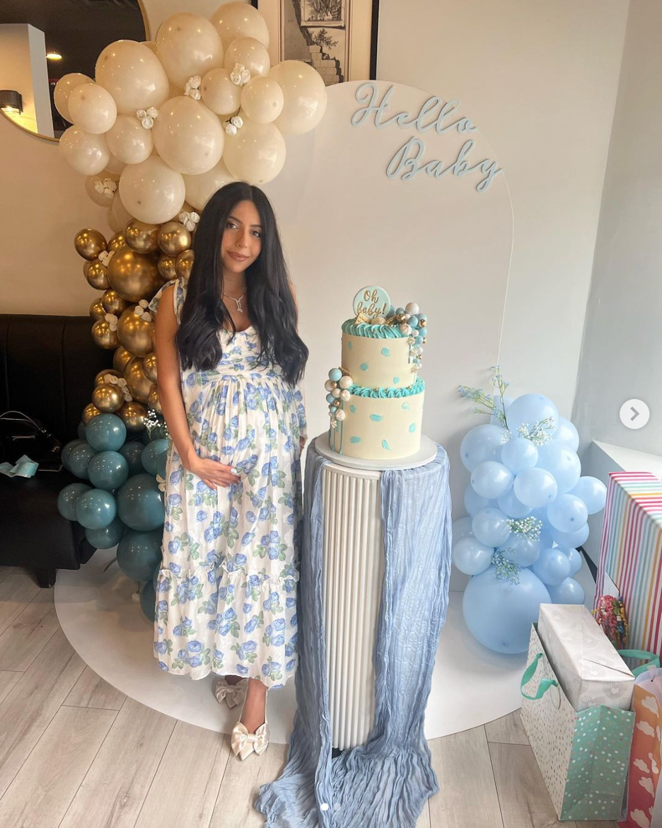 A young woman in a floral dress stands next to a two-tiered cake with blue and white decorations, celebrating a baby shower. There are balloon arrangements in white, gold, and teal hues, and a sign that reads "Hello Baby" on the wall behind her.