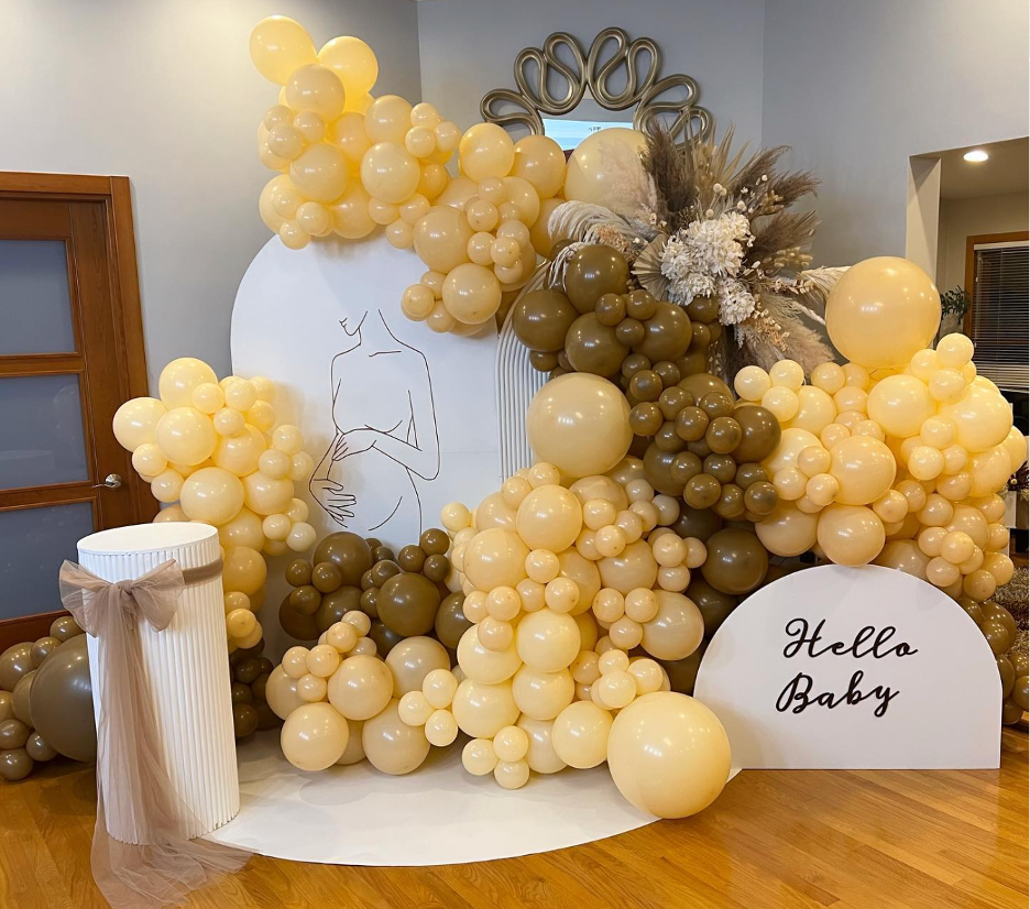 Decorative balloon arrangement with beige, cream, and brown balloons, floral accents, and a sign that says "Hello Baby" for a baby shower.