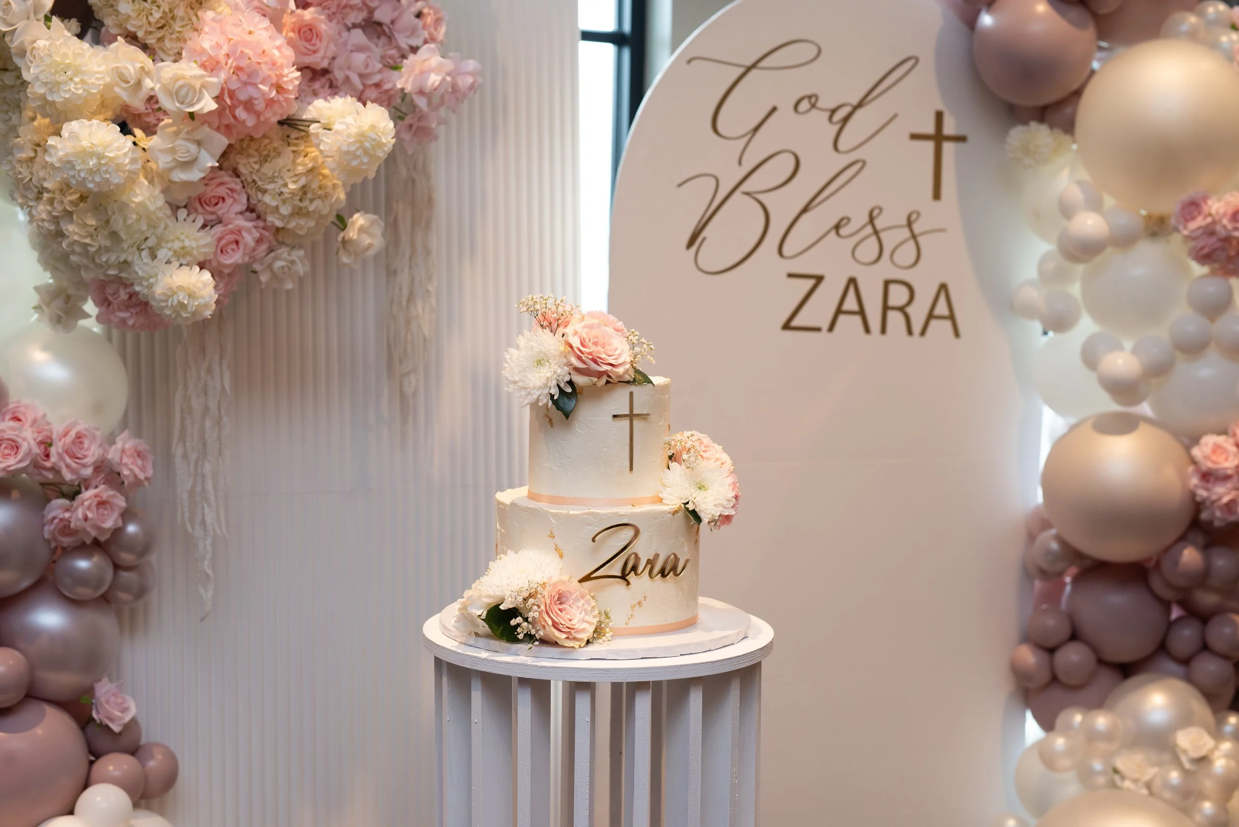 A decorated celebration display featuring a two-tier white cake with pink and white flowers and gold accents, a large sign with the message 'God Bless Zara' in gold lettering, surrounded by pink and white balloons and floral arrangements.