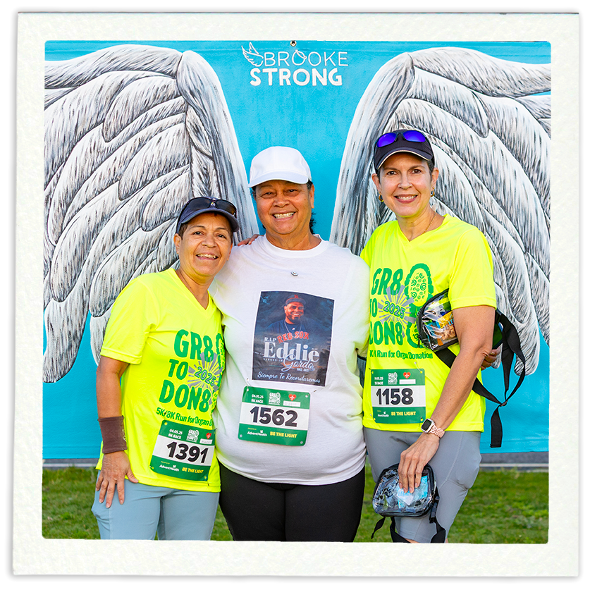 Three women standing together at a charity run event, with a large banner displaying angel wings and the words 'Brooke Strong' behind them. The women are smiling, wearing bright yellow event shirts and race bibs, with the middle woman wearing a white hat and the women on either side wearing caps.
