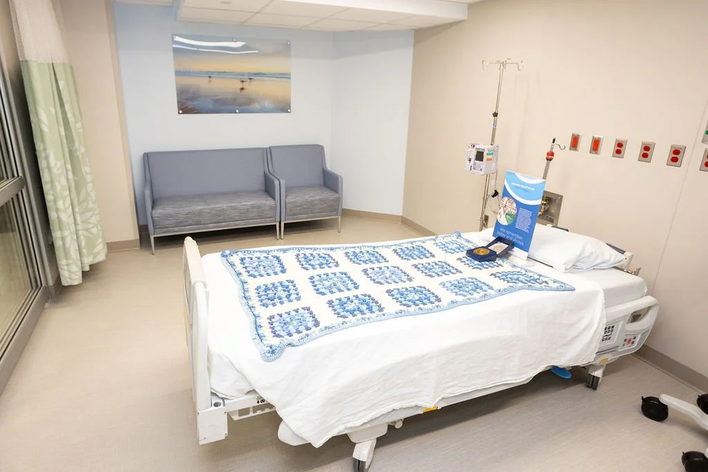 Hospital room with a bed, a quilt with blue patterns, a pillow, an IV stand, a small table with brochures, a gray couch, a window with curtains, and a painting of a beach scene on the wall.