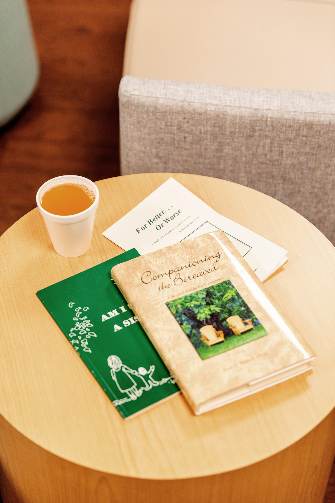 Books, a cup of tea or coffee, and a pamphlet are on a round wooden table near a beige upholstered chair.