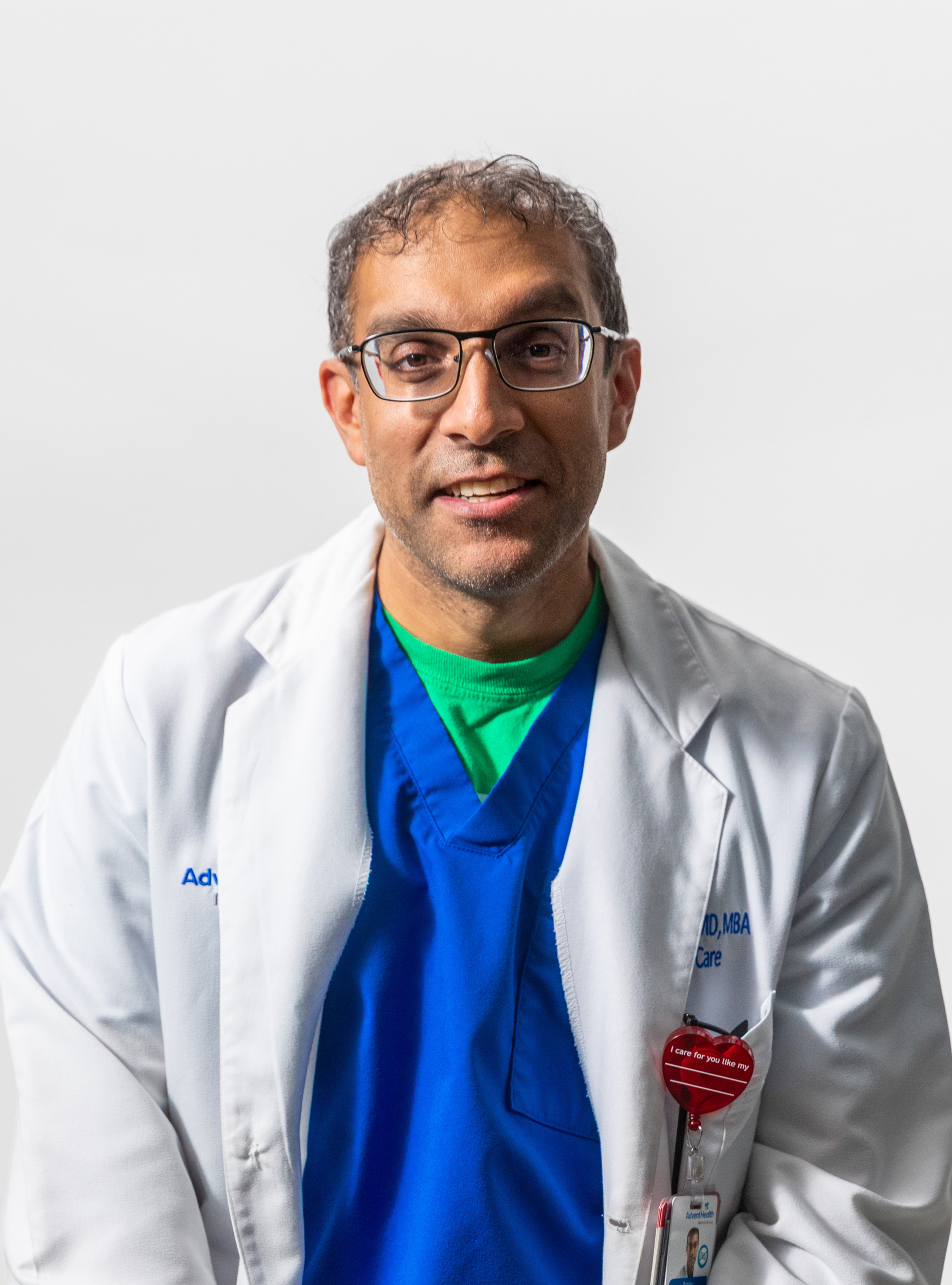 A smiling male doctor wearing glasses, a white lab coat, blue scrubs, and a green shirt, standing against a plain white background.