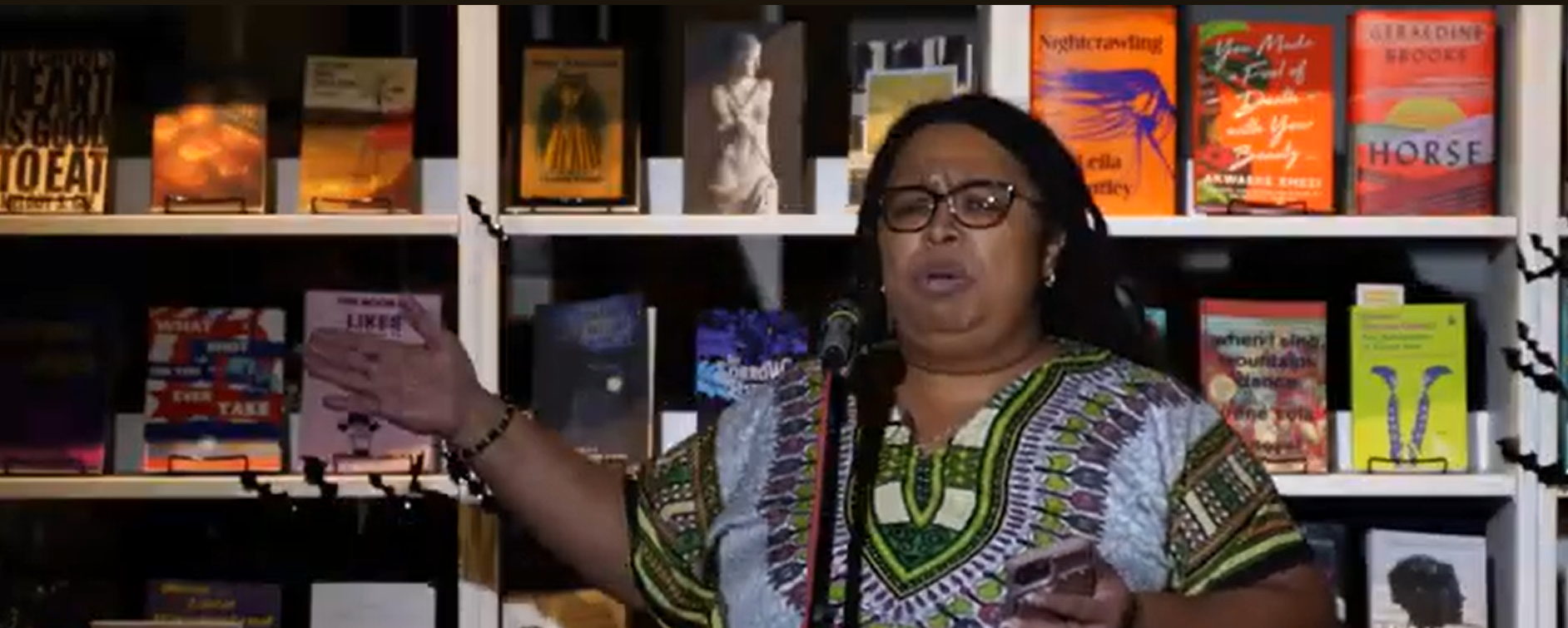 Woman speaking at a microphone in front of a bookshelf filled with colorful books and a framed picture.