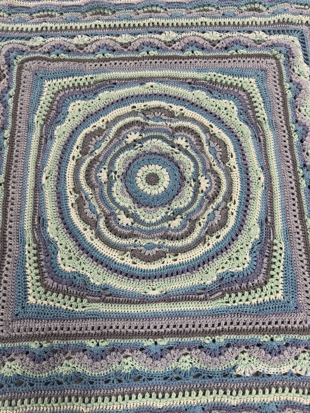 A colorful, round crochet blanket with concentric rings and intricate patterns in shades of blue, gray, cream, and purple.
