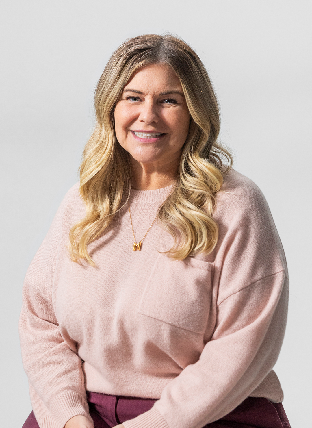 A woman with long wavy blonde hair wearing a light pink sweater, a gold necklace, and smiling at the camera against a light gray background.