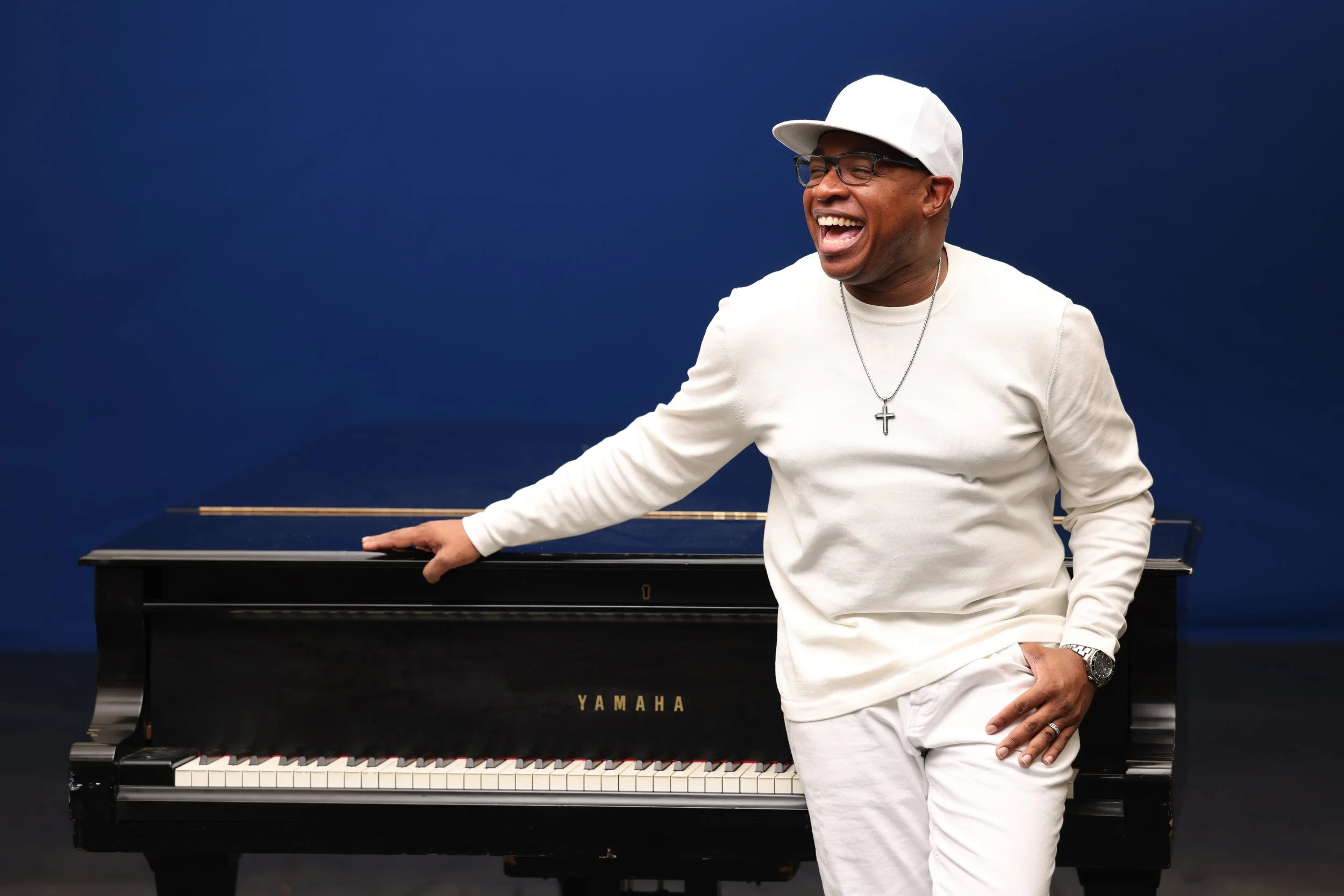 A man dressed in white, wearing glasses, a white cap, and a cross necklace, smiling and standing next to a black Yamaha piano against a dark blue background.