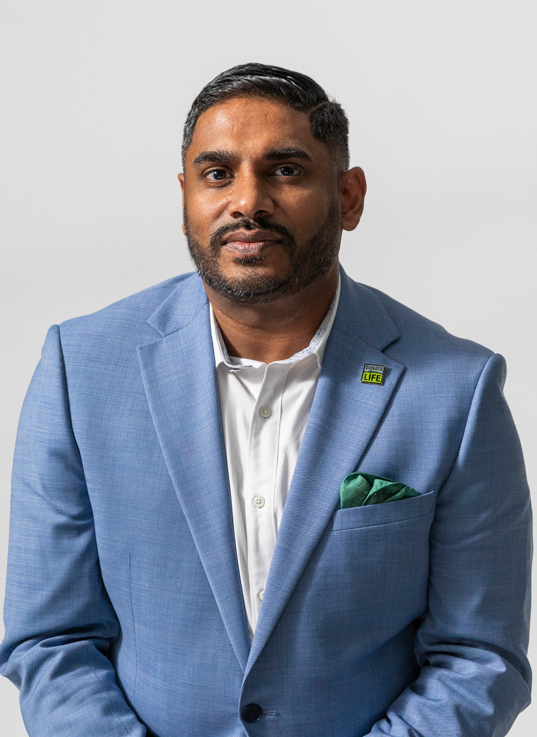 A man in a light blue suit with a green pocket square, white shirt, and a donation badge on his lapel, standing against a plain light gray background.