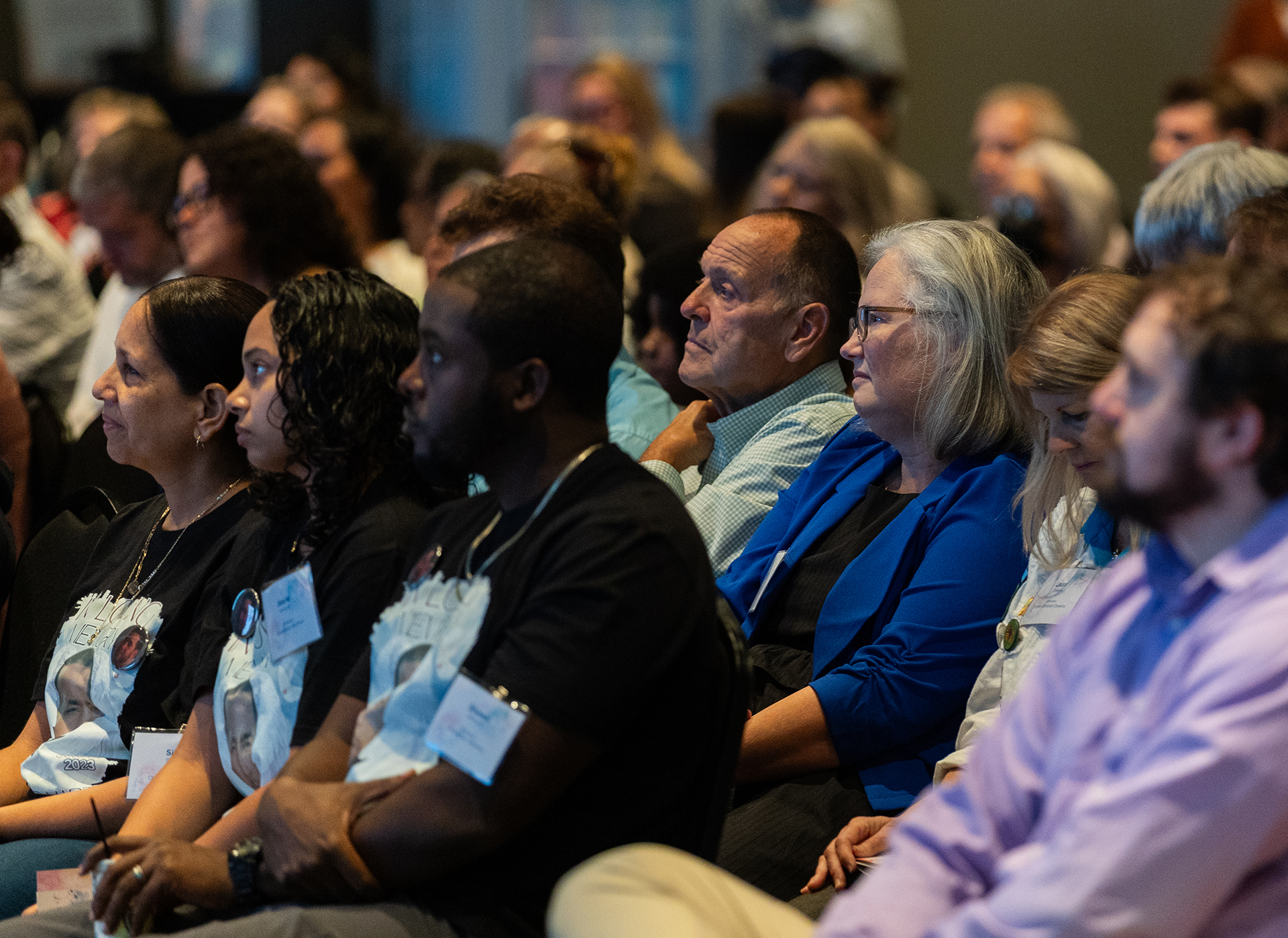 Audience members sitting attentively during a conference or seminar, with some wearing name tags and event badges.