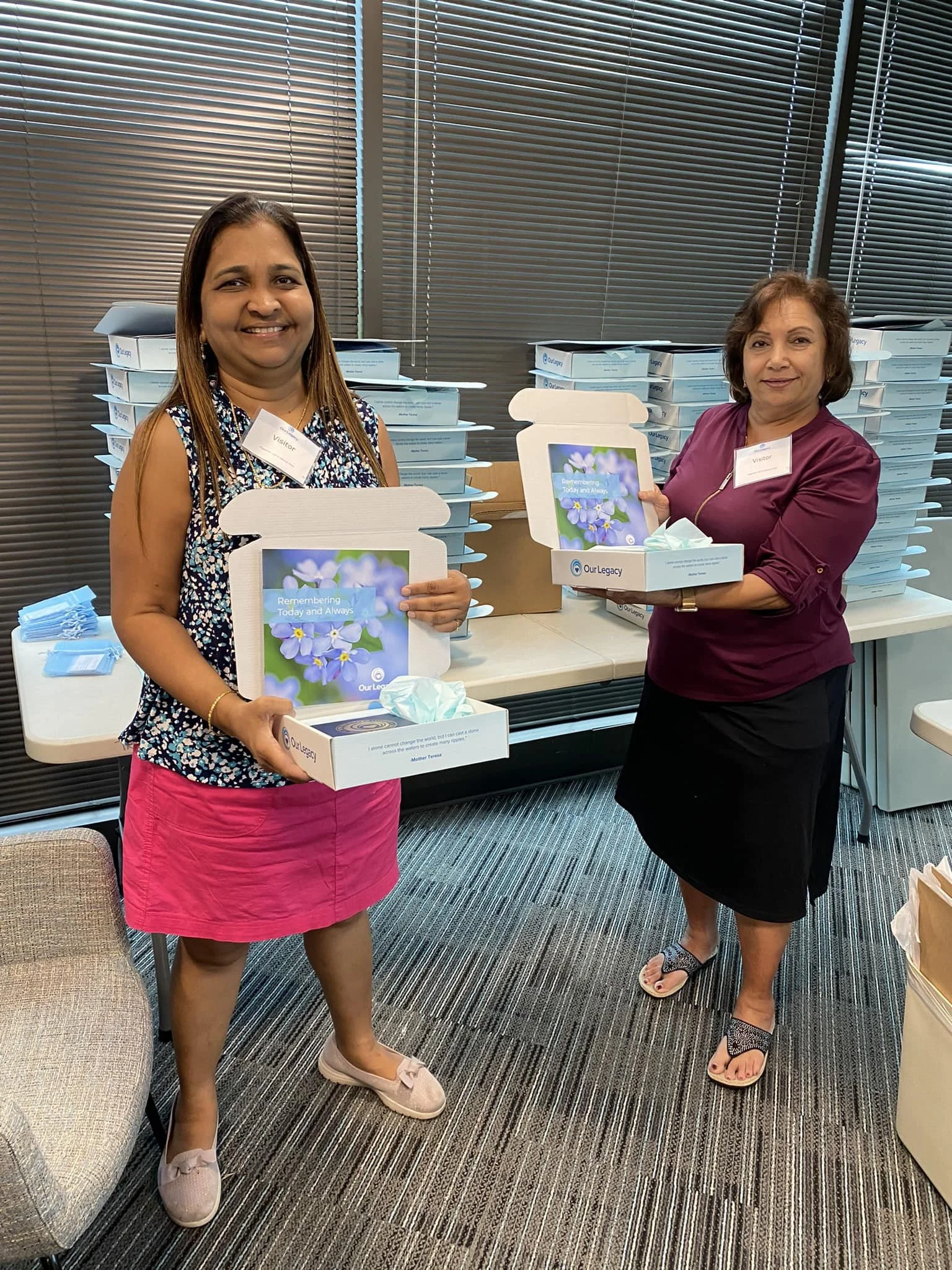Two women standing indoors holding open boxes with a floral design and tissues inside, with stacked boxes in the background. They are smiling and wearing name tags.