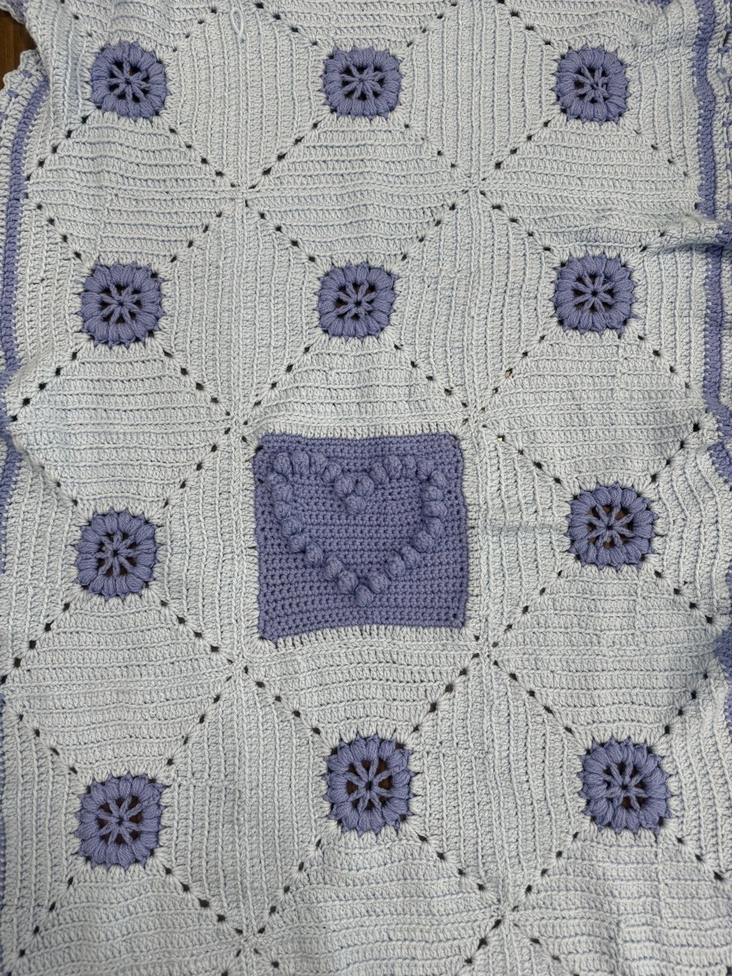A crocheted blanket featuring a pattern of purple flowers with black centers and a purple heart inside a purple square at the center, all stitched on a cream background.