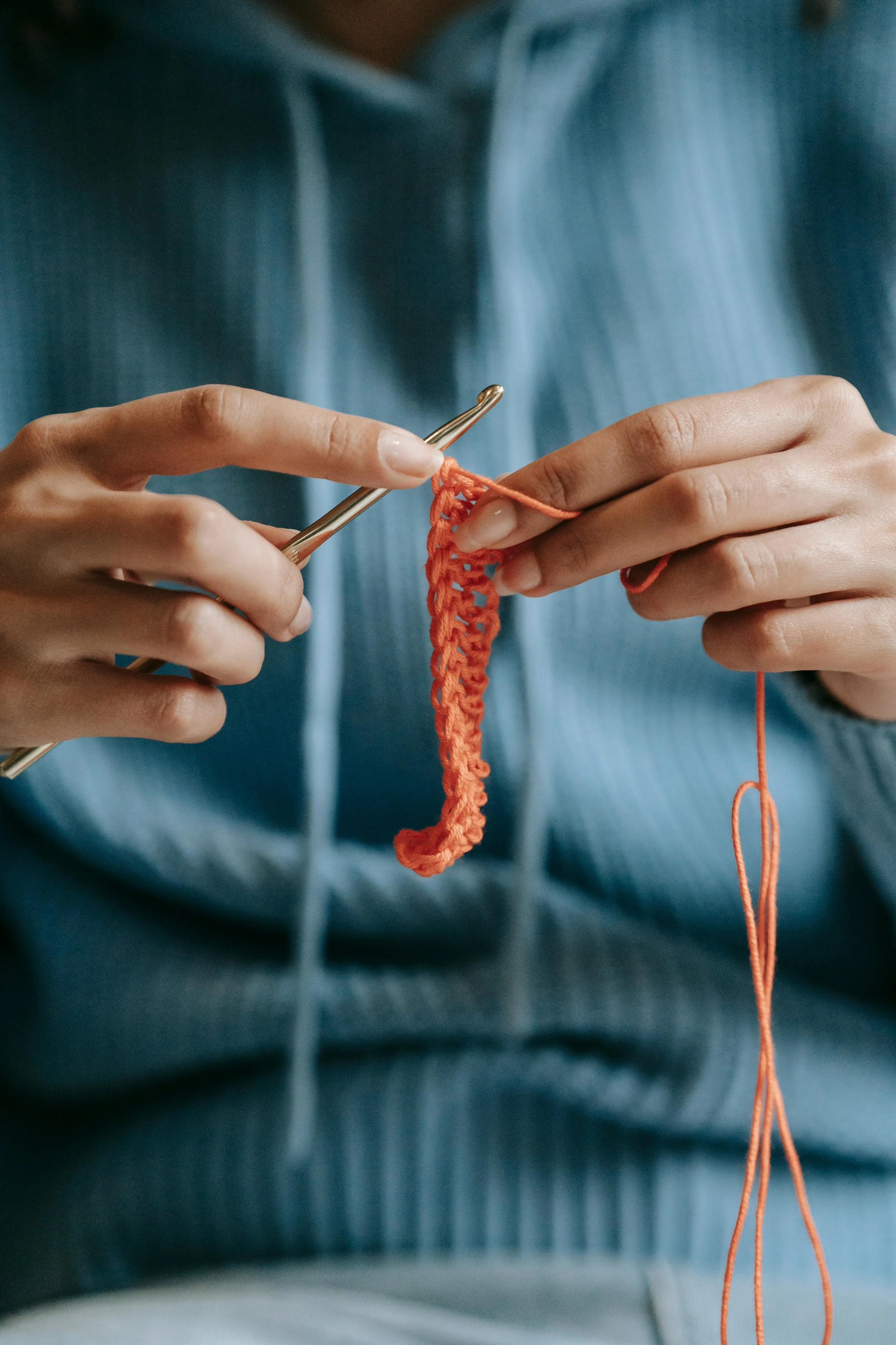 A person wearing a blue hoodie is crocheting with orange yarn and a metal crochet hook.
