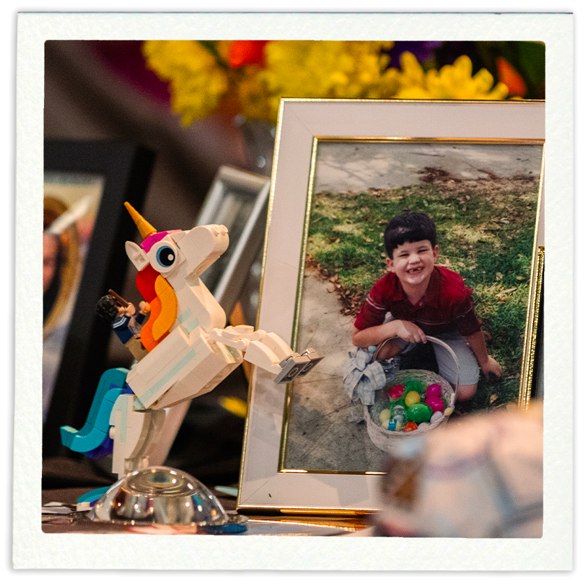 Photograph of a framed picture of a smiling young boy outdoors holding a basket of colorful plastic eggs, with a LEGO unicorn toy and other framed photographs nearby.
