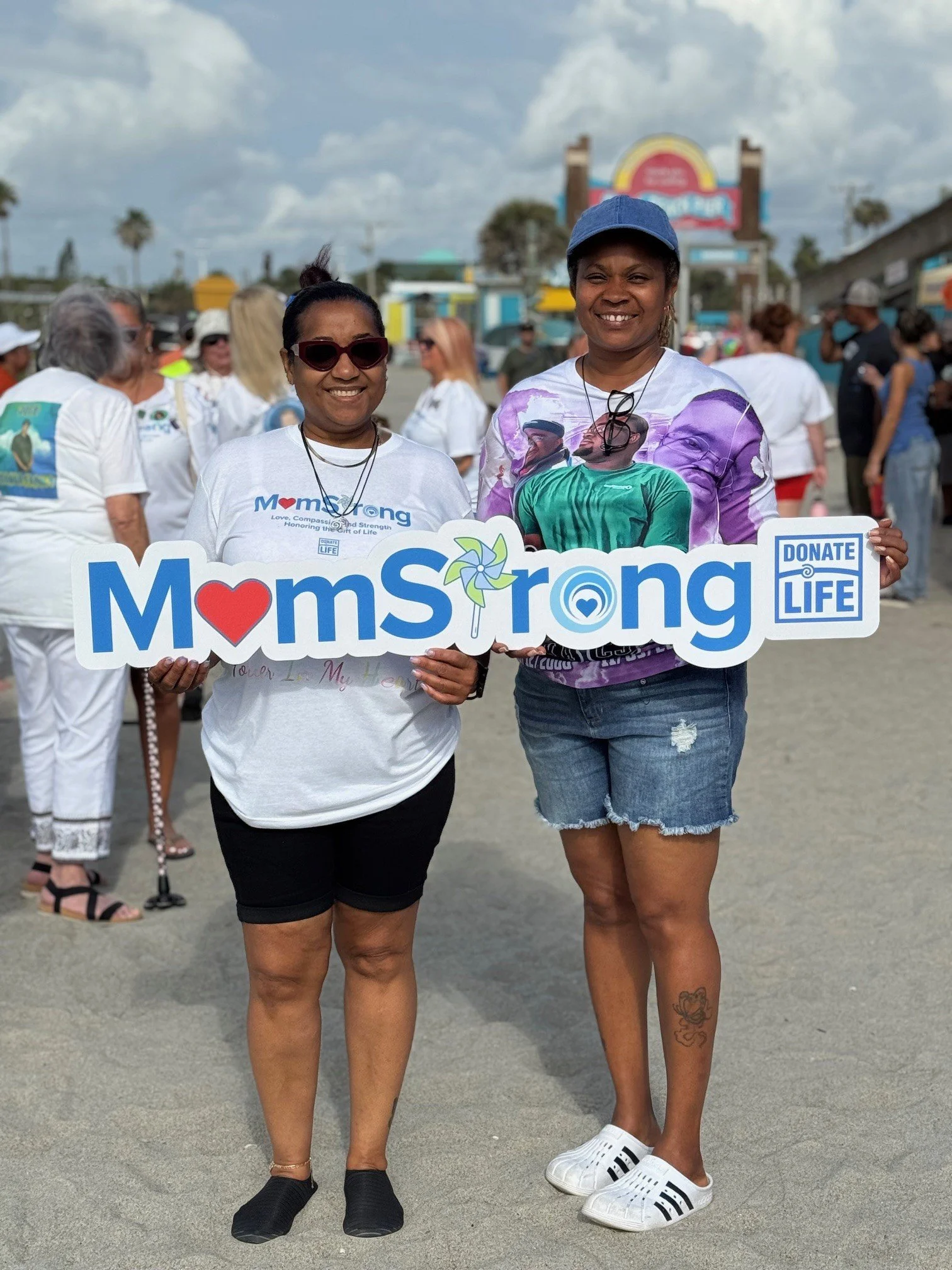 Two women smiling and holding a sign that reads "MomStrong" at an outdoor community event, with a crowd of people in the background.