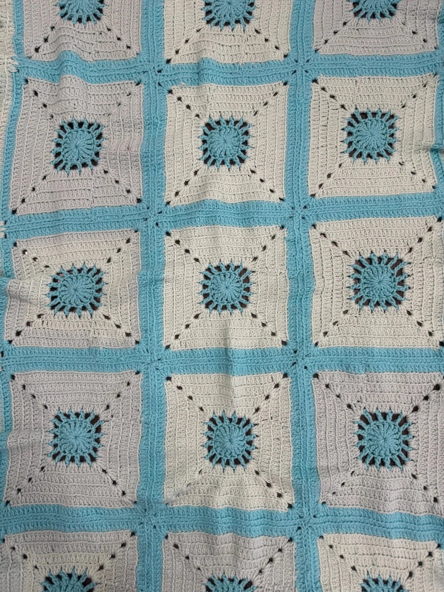 A crochet blanket with a pattern of cream square blocks, each centered with a blue circle and black dotted details, surrounded by blue borders.