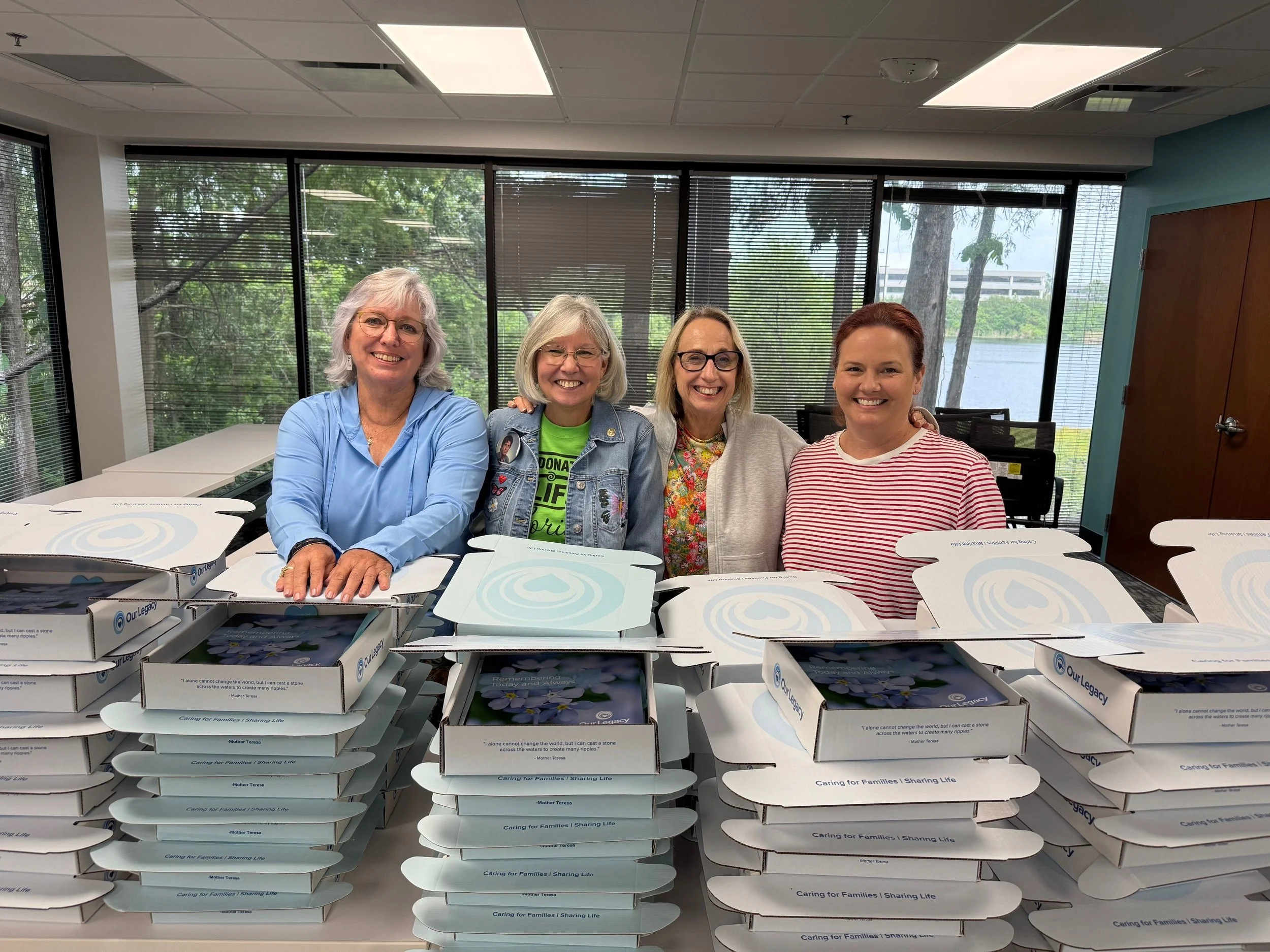 Four women standing behind a table with multiple closed white boxes, smiling. Large windows overlooking trees and water in the background.