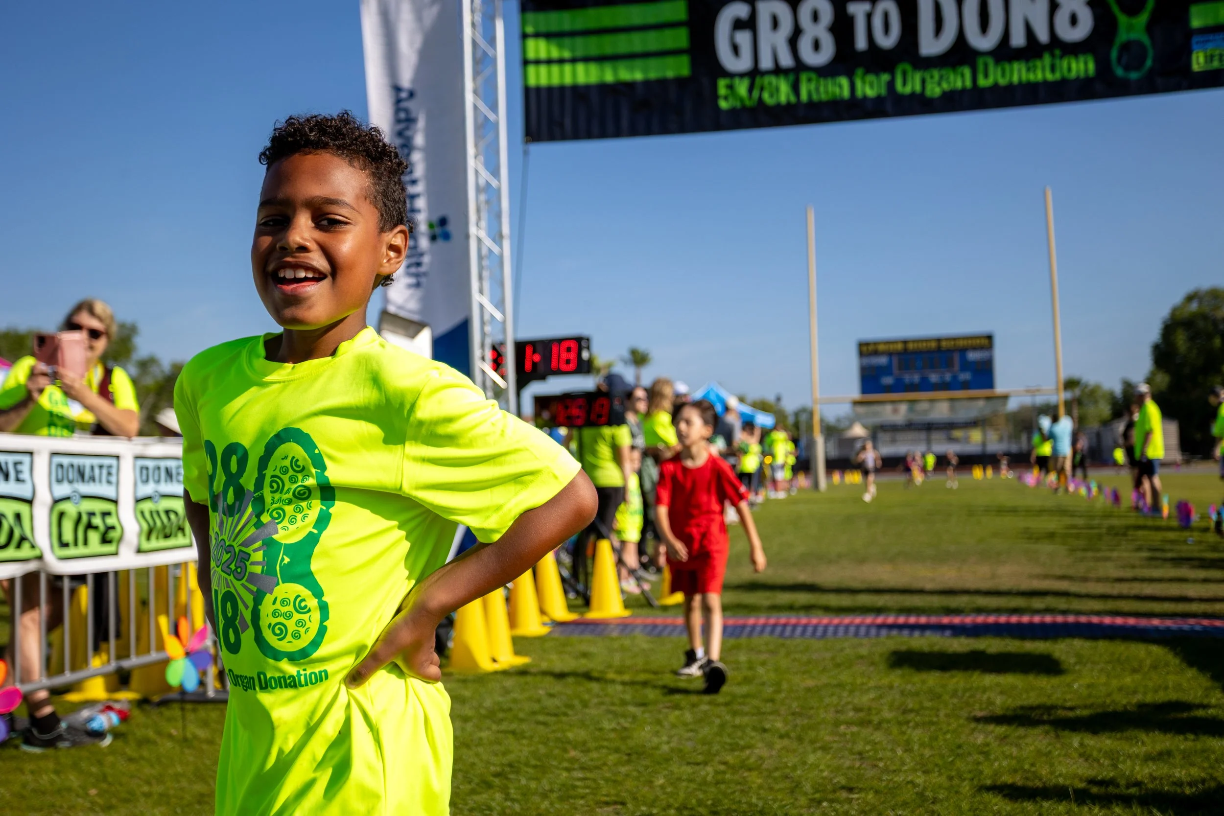 A young boy in a bright yellow T-shirt with a marathon logo, smiling at the finish line of a charity run, with other participants and spectators in the background on a sunny day.