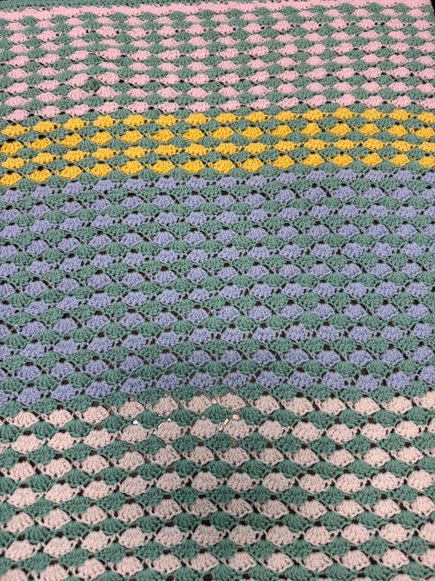 Close-up view of a multicolored crocheted blanket with a pattern of repeating shells in pastel pink, yellow, purple, and green colors.