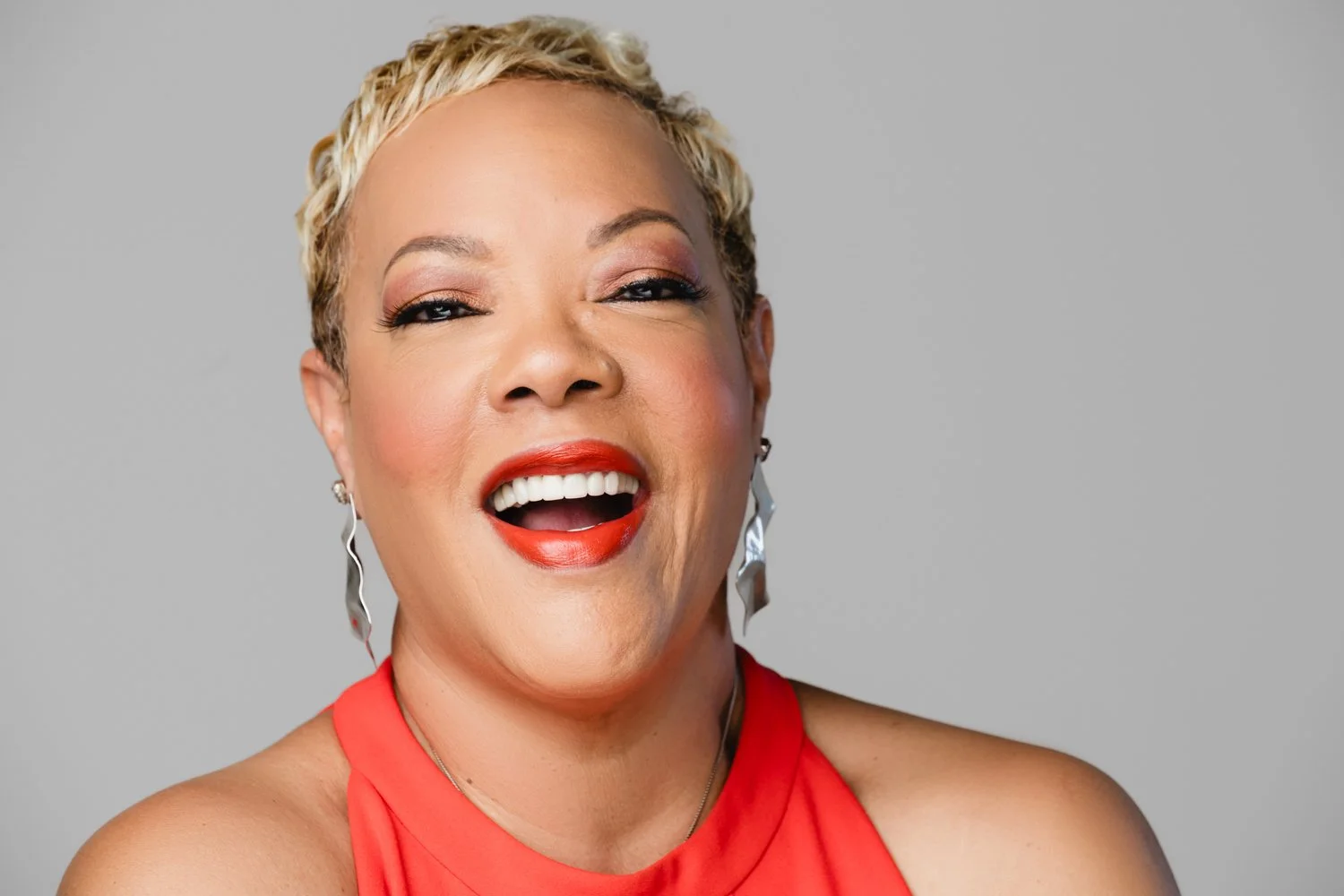 A smiling woman with short, curly blonde hair, wearing a red sleeveless top, bold red lipstick, and large silver earrings, posing against a plain gray background.
