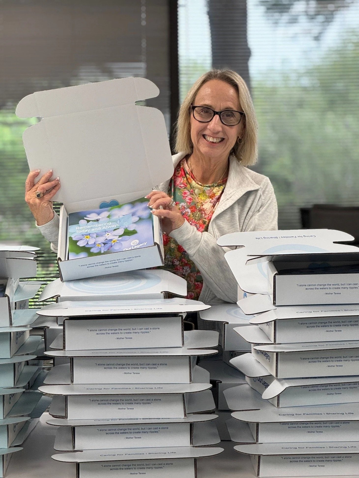 A smiling woman with glasses holding an open box with a card inside, surrounded by stacked boxes on a table.