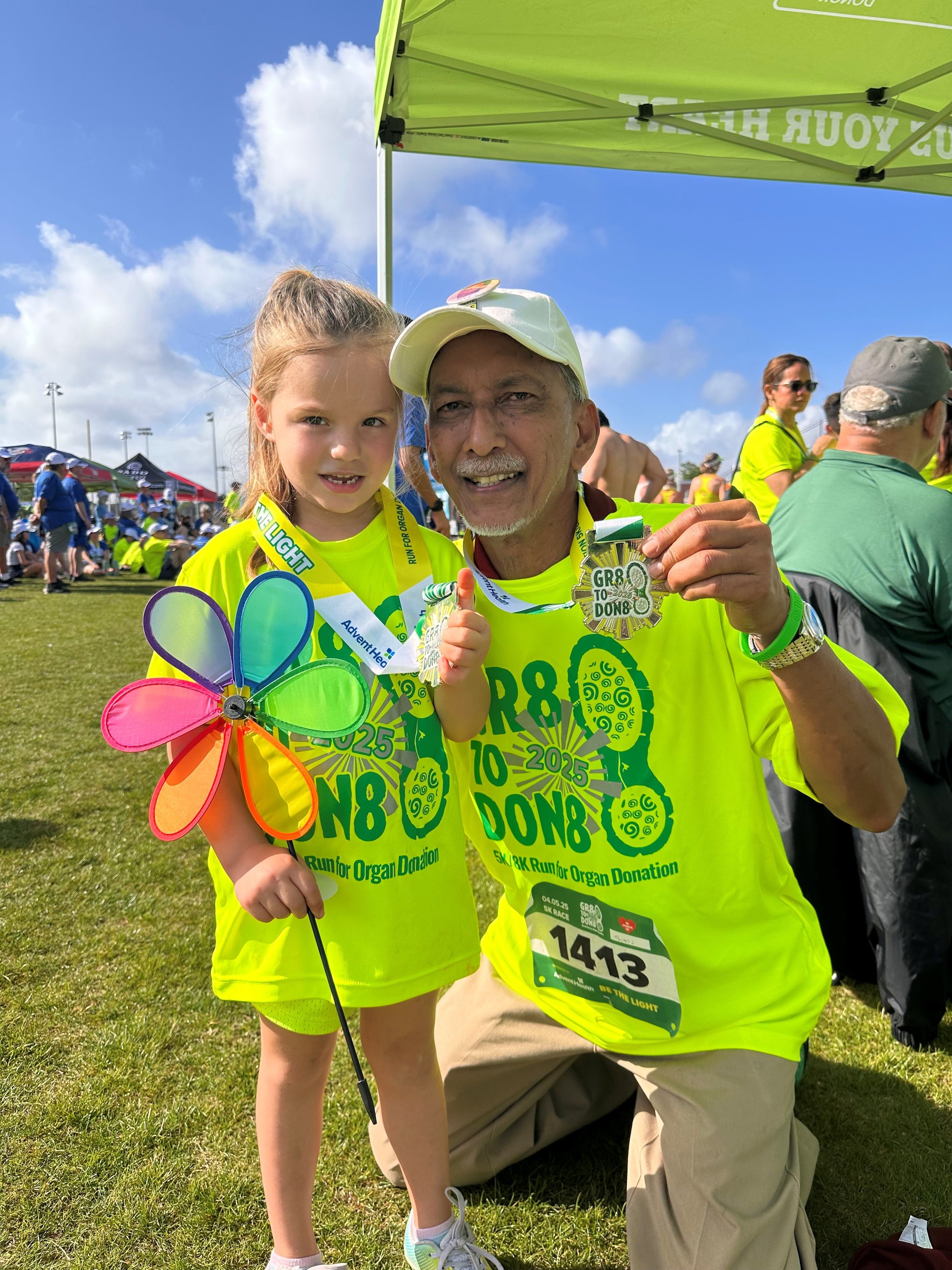 An elderly man and a young girl at an outdoor event celebrating a charity run. They are both wearing bright yellow-green t-shirts with the event details and medals around their necks. The man is holding up his medal and smiling, while the girl is hol