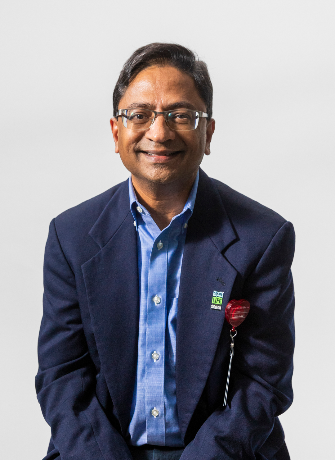A man in glasses wearing a dark blue blazer and light blue shirt, smiling, with a white background.
