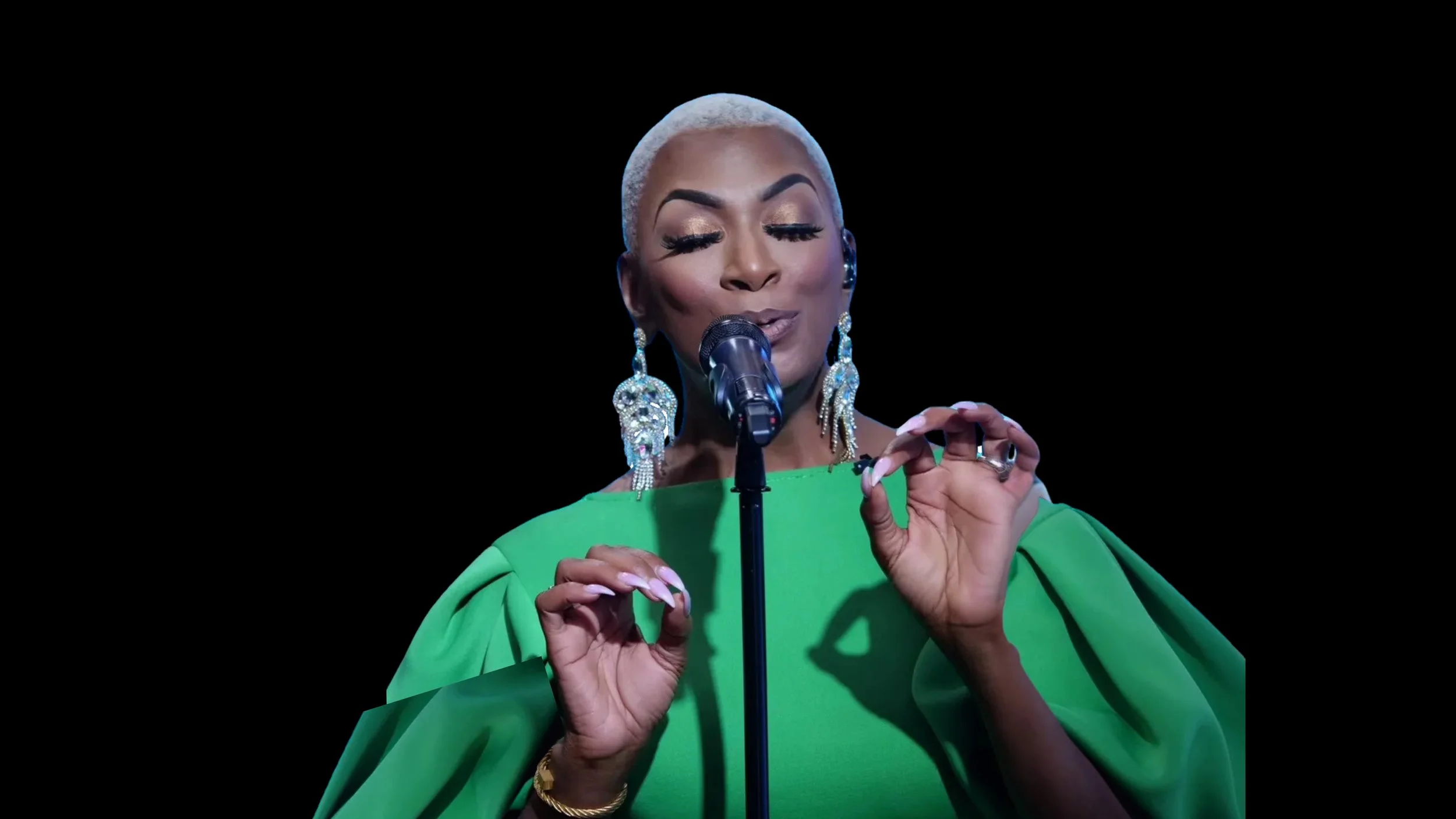 A woman singing into a microphone on a black background. She has short, platinum blonde hair, dramatic earrings, and wears a vibrant green dress.