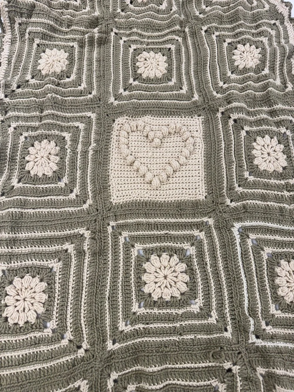 A crocheted blanket featuring a pattern of squares with floral motifs and a heart design in the center square, in shades of gray and white.