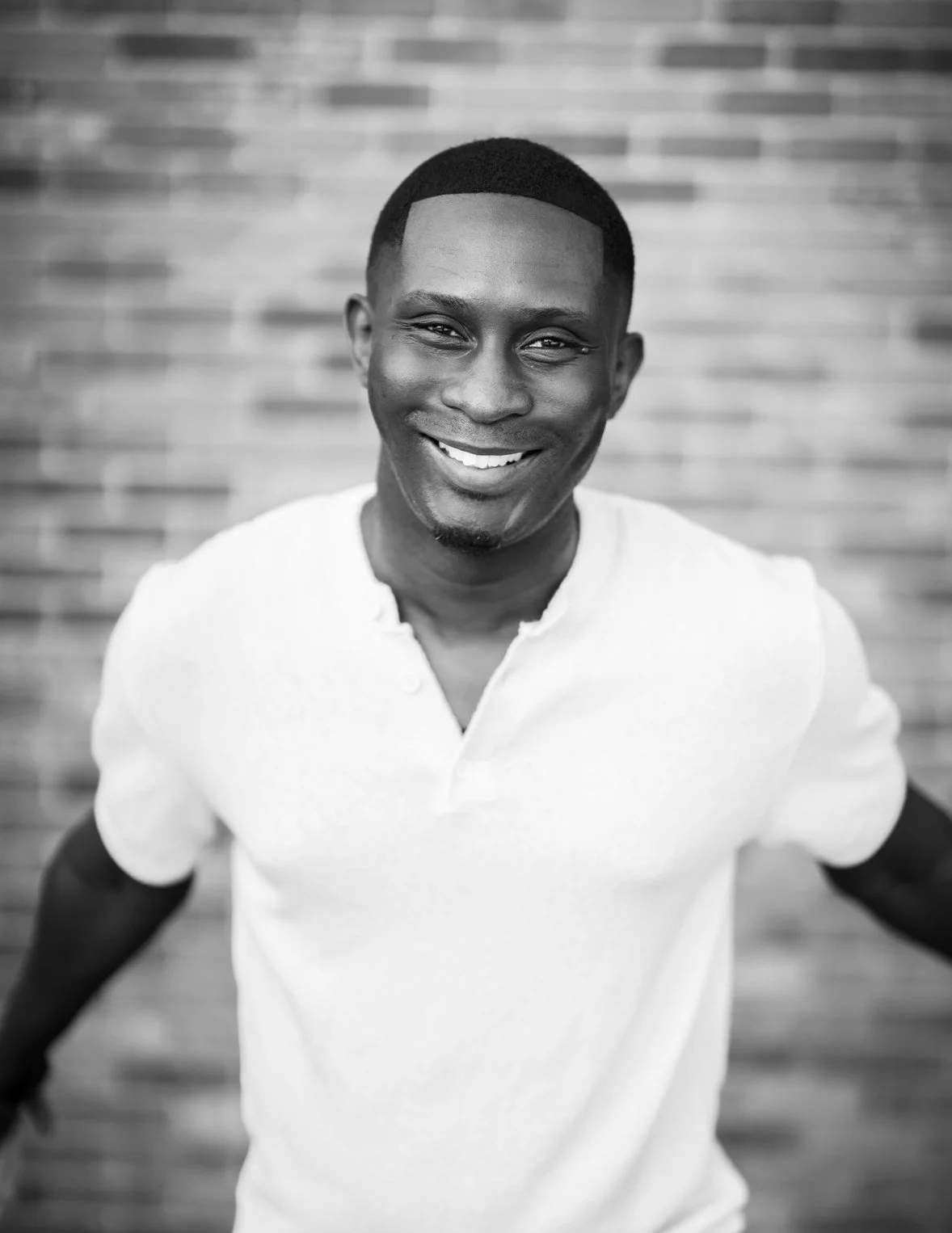 A smiling man in a white shirt standing in front of a brick wall.