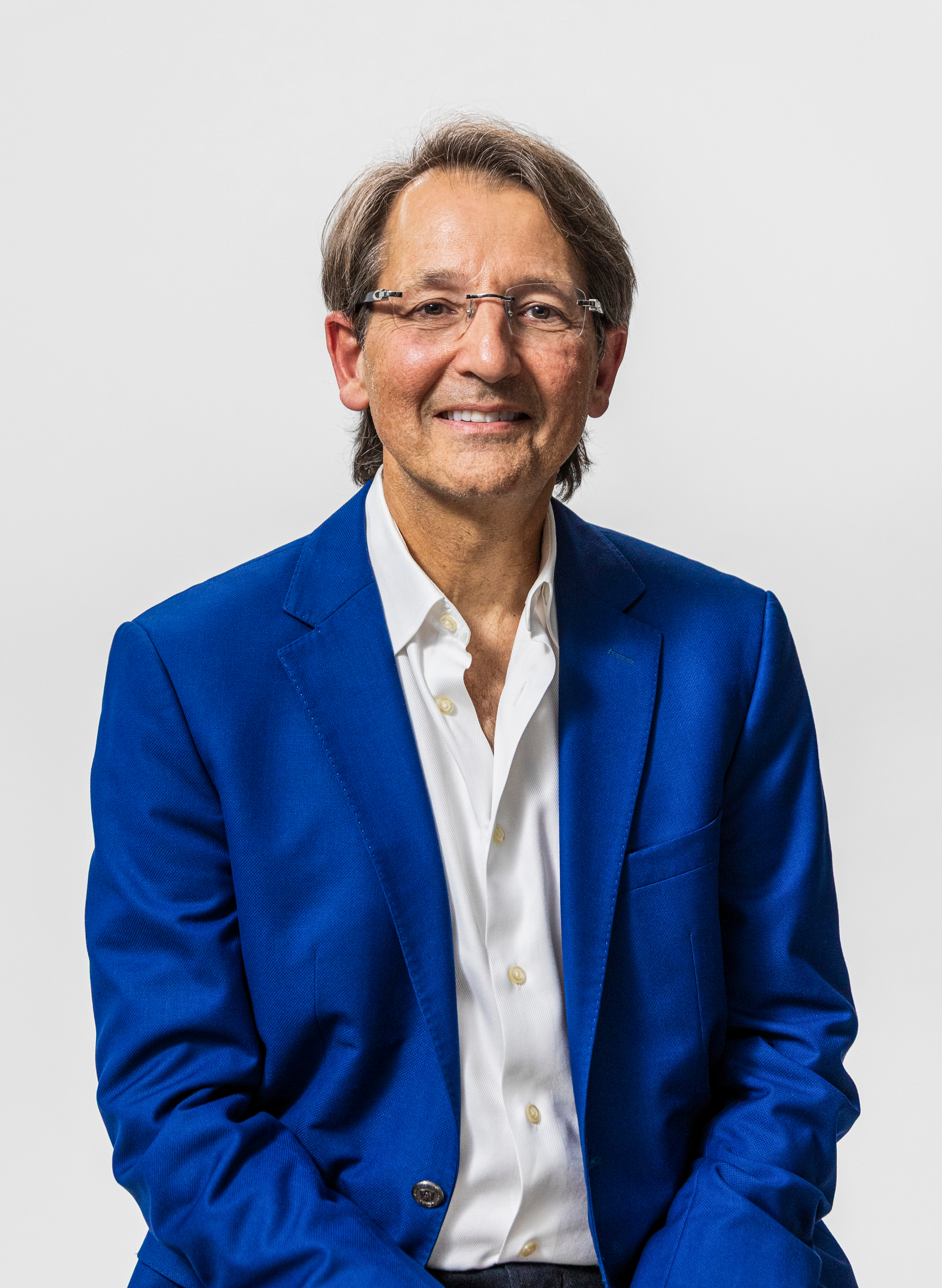 A man in a blue suit jacket, white button-up shirt, and glasses smiling against a plain white background.