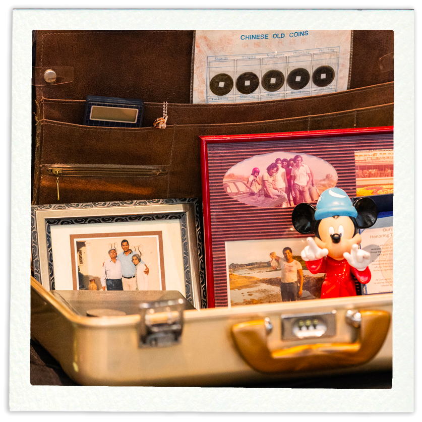 Collection of personal and decorative items in a suitcase, including framed photographs, a Mickey Mouse figurine, a framed collage, a Chinese old coins sheet, a calculator, and a keychain.