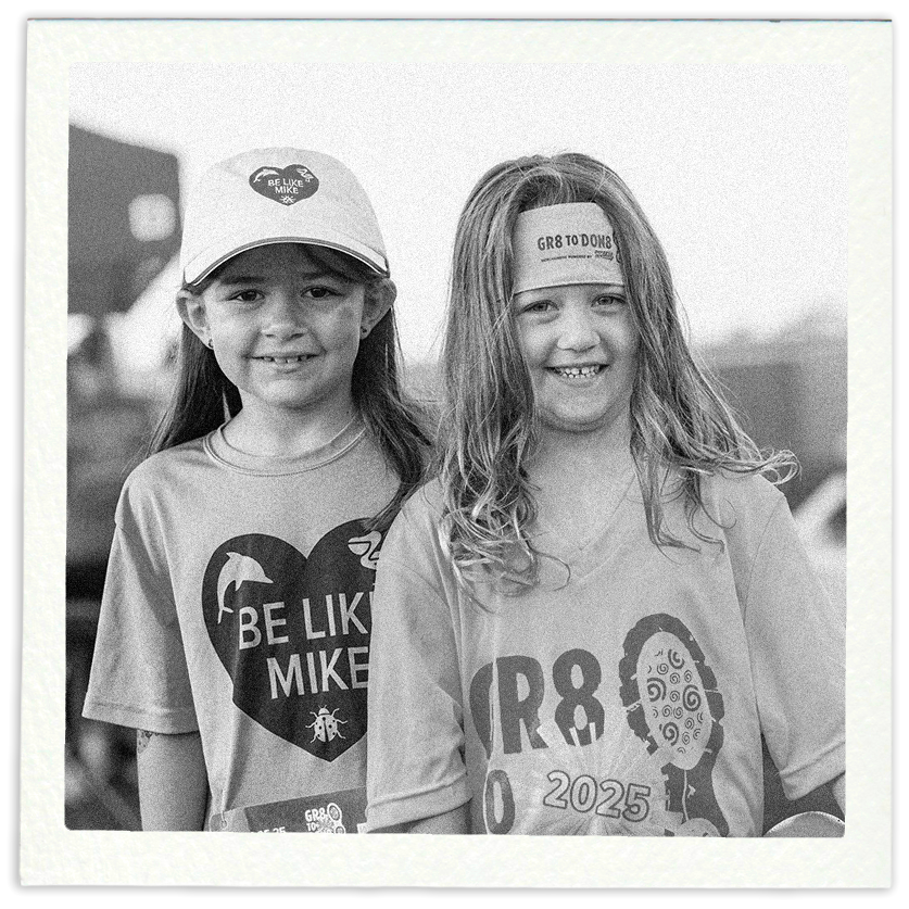Two young girls smiling outdoors, wearing casual T-shirts and one wearing a baseball cap and the other a headband. The girl on the left has a cap that says "Be Like Mike" and a T-shirt with the same message, while the girl on the right has a headband and a T-shirt with graphic designs and text related to the year 2025.