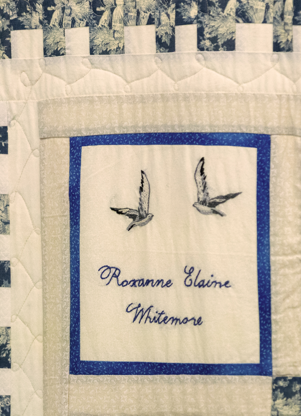 Close-up of a quilt featuring two embroidered hummingbirds and a personalized name label, "Roxanne Elaine Whitmore," with a blue embroidered border and patterned fabric in the background.