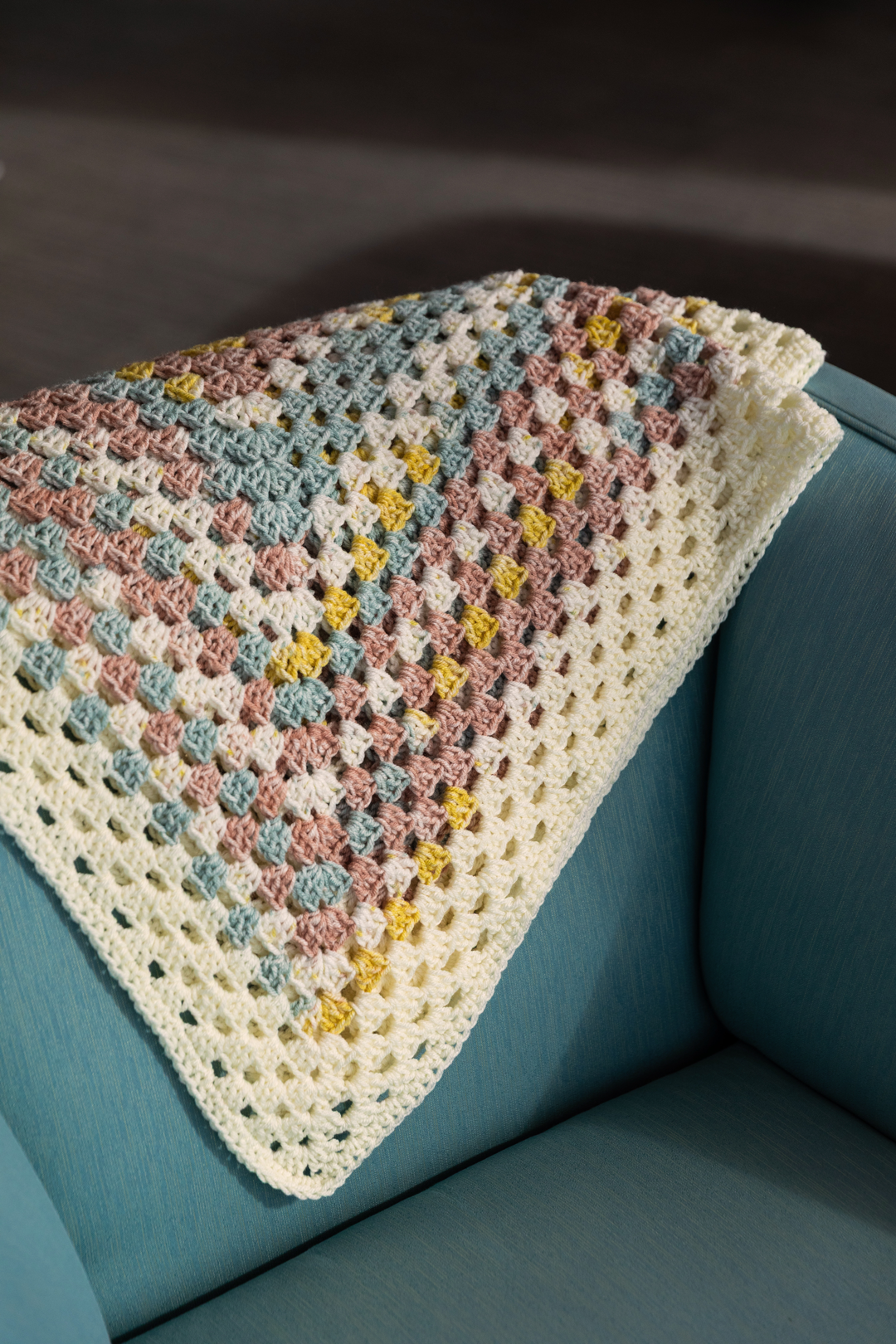 A multicolored crocheted blanket with pastel shades of pink, blue, yellow, and cream, draped over a teal armchair.
