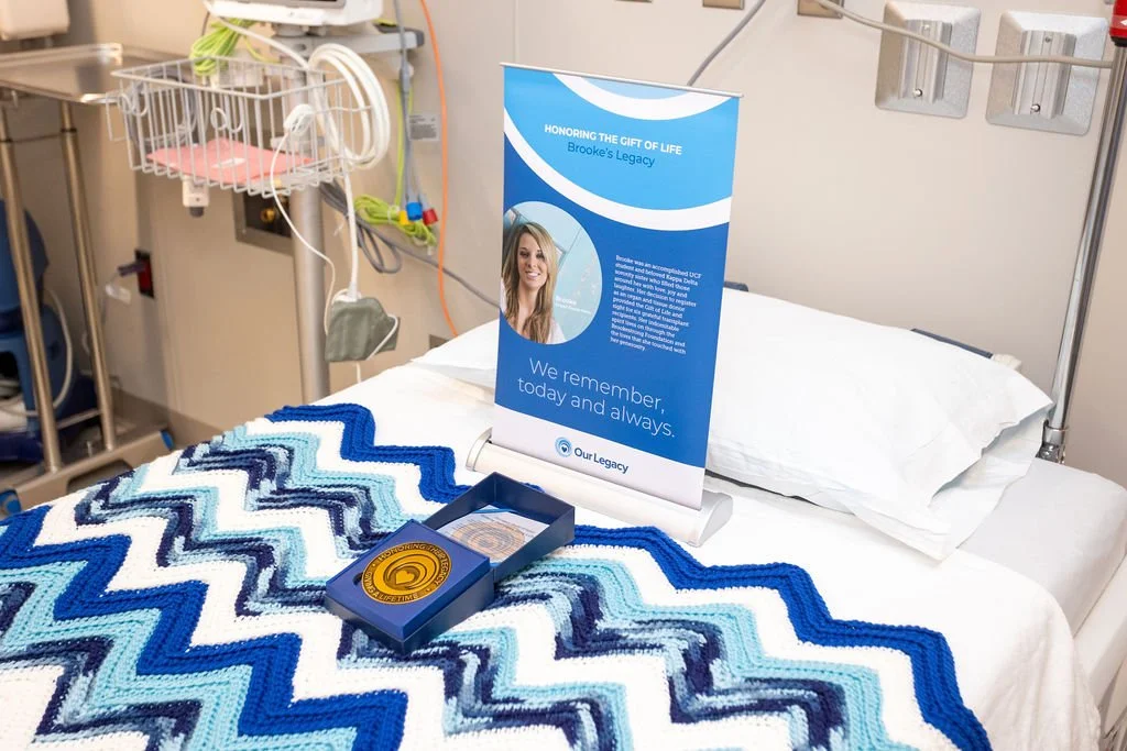 Hospital bed with a blue zigzag blanket, a flower and memorial display for Brooke, with a sign honoring her legacy.
