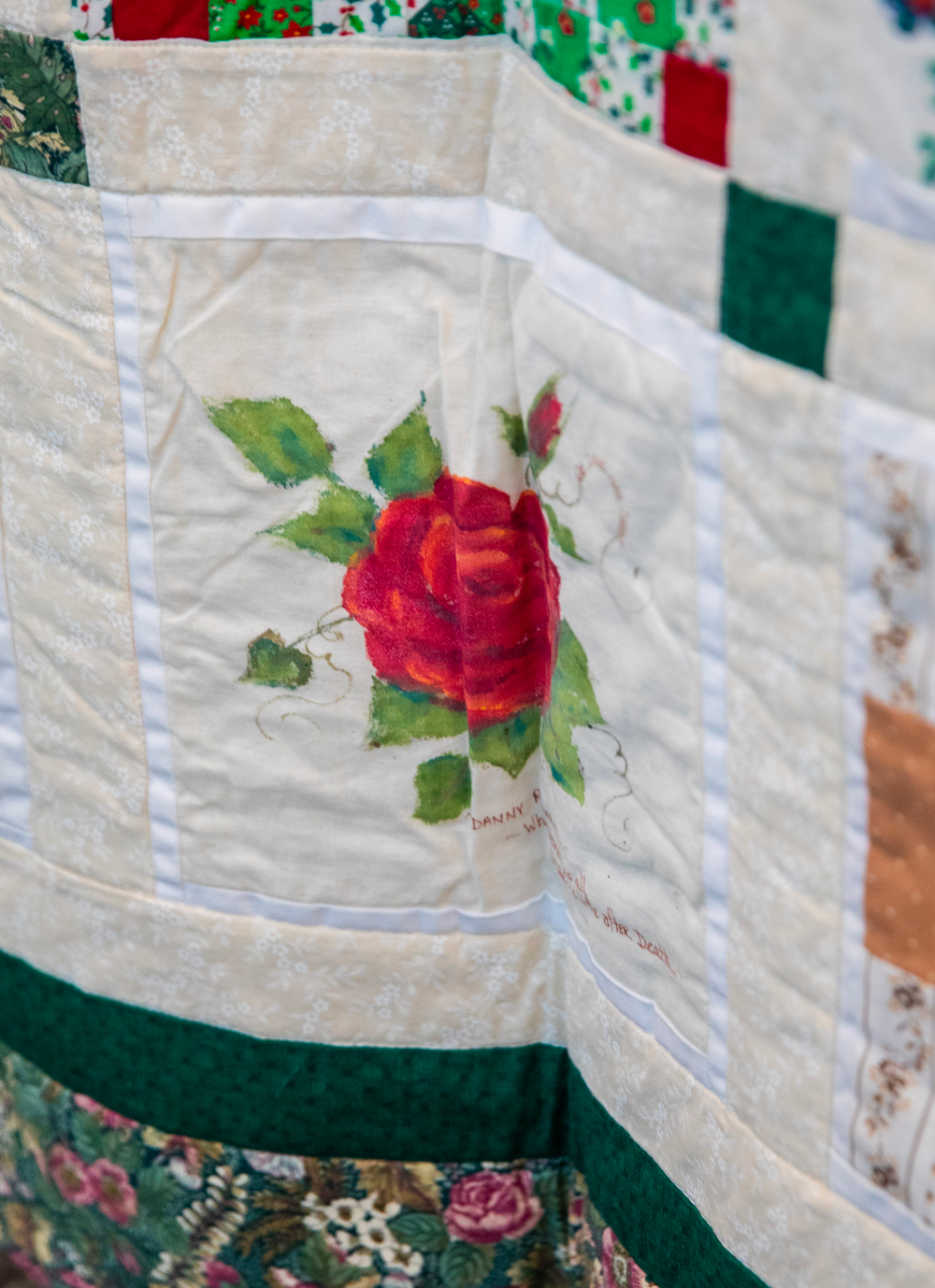 Close-up of a quilt with a central panel featuring a painted red rose with green leaves and tendrils, surrounded by multiple patterned fabric sections.