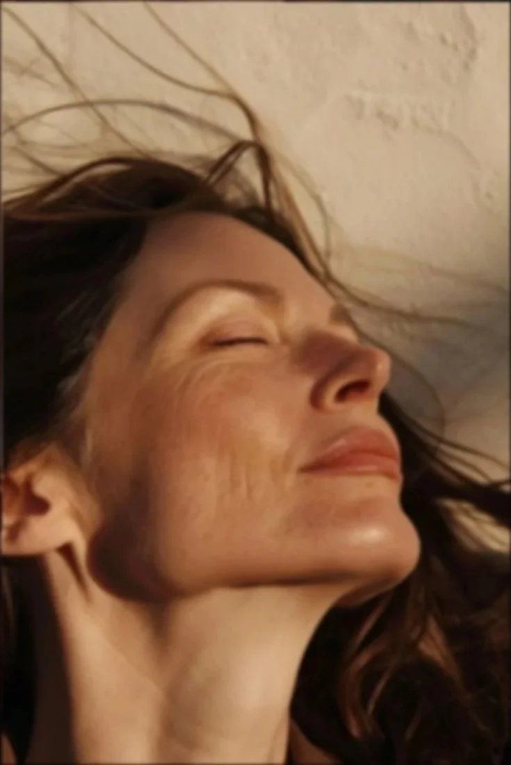 A woman with long dark hair and light makeup is smiling with eyes closed, lying on her side against a textured wall.